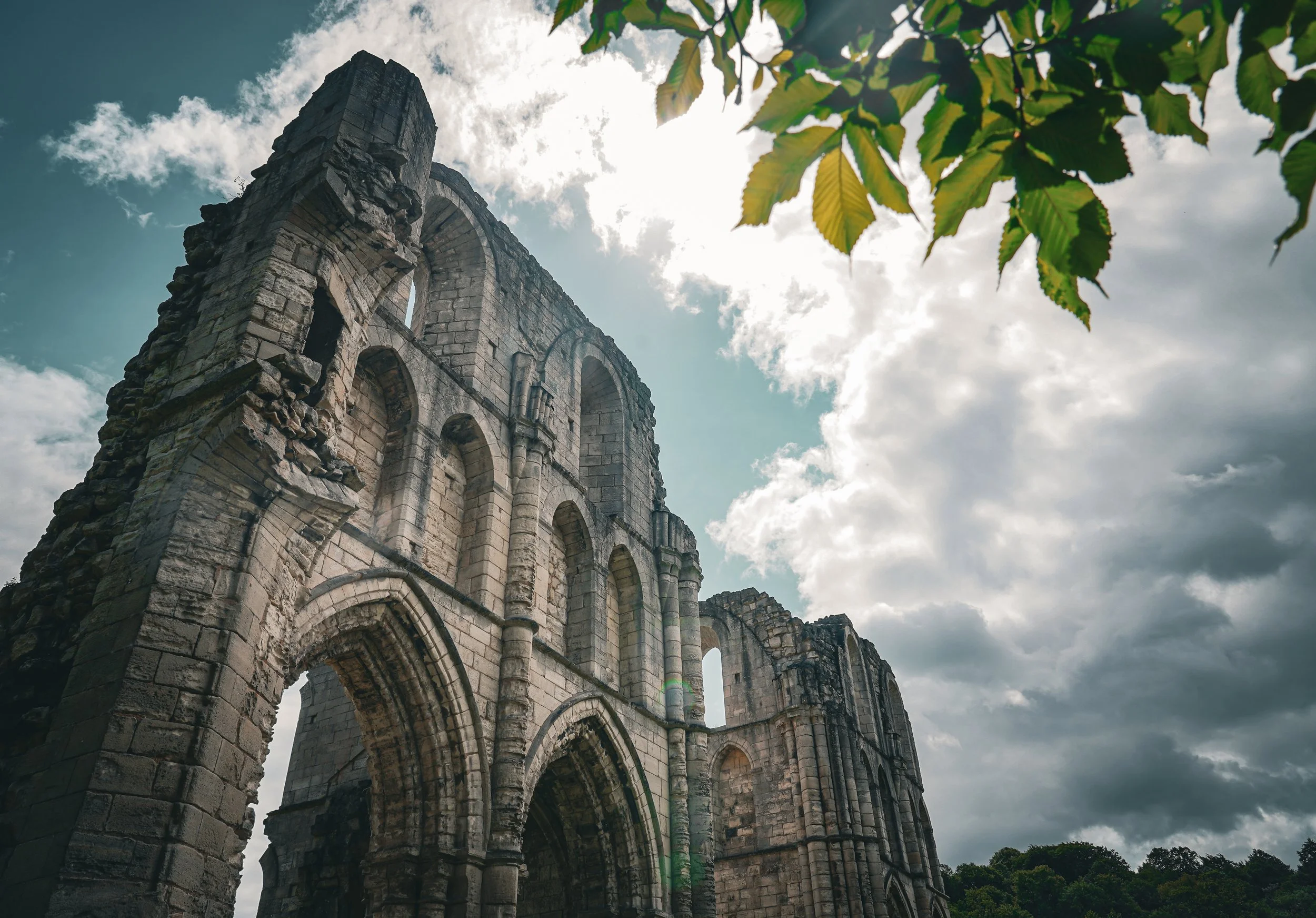 Ancient stone ruins of an arched structure under a cloudy sky with green leaves in the upper right corner.