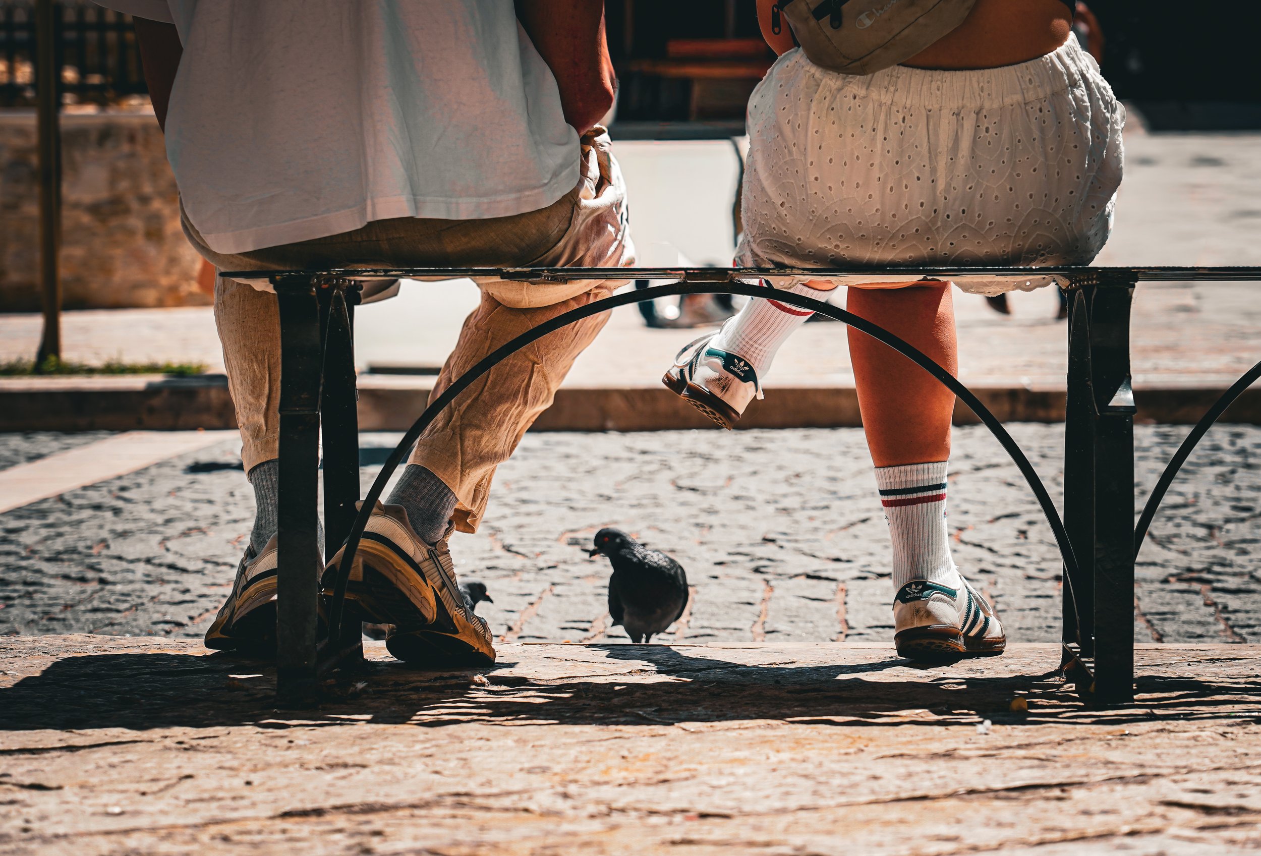 Two people sitting on a bench with a pigeon underneath them on a cobblestone surface, view from beneath.