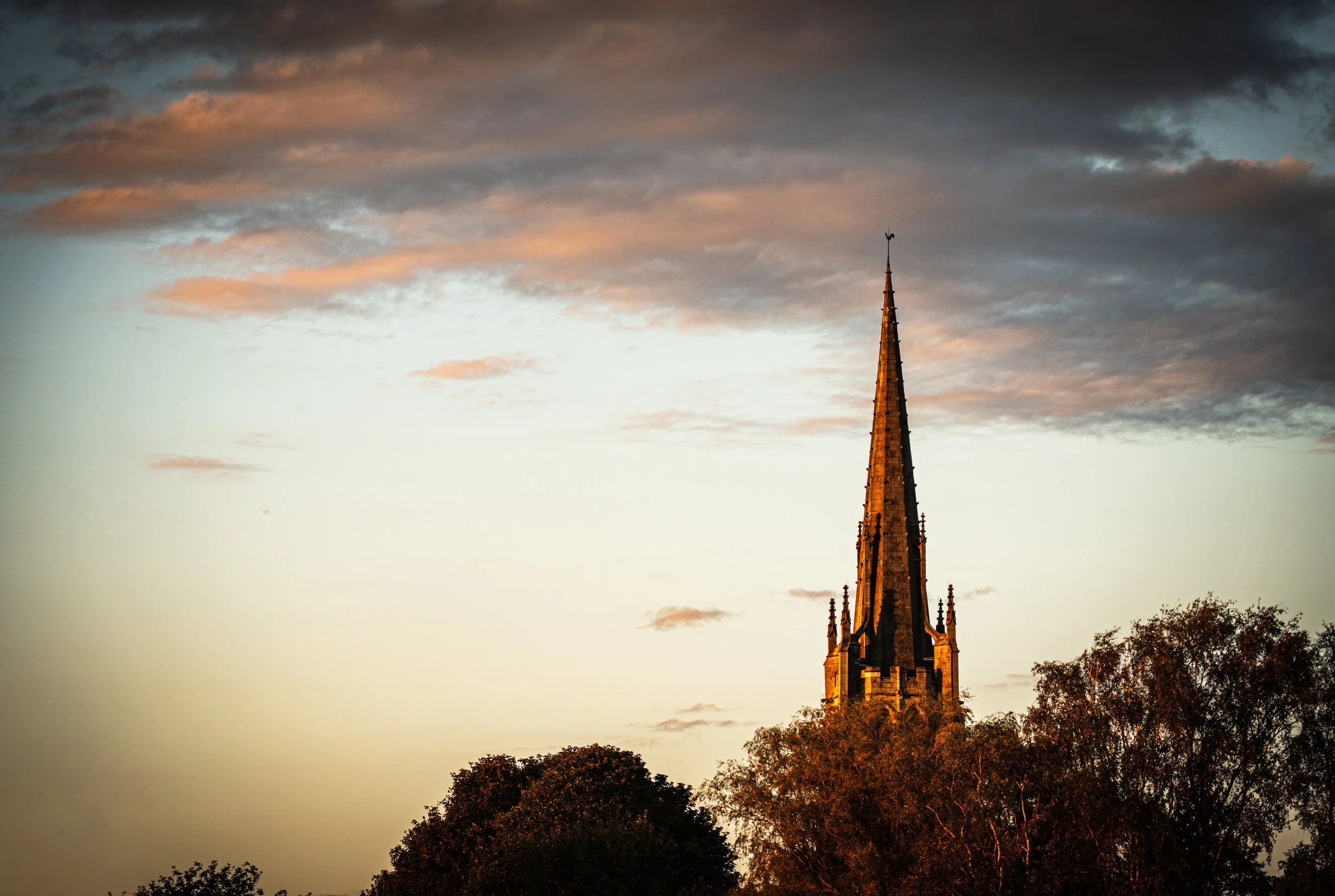 A tall church steeple rising above tree line during sunset with orange and dark clouds in the sky.