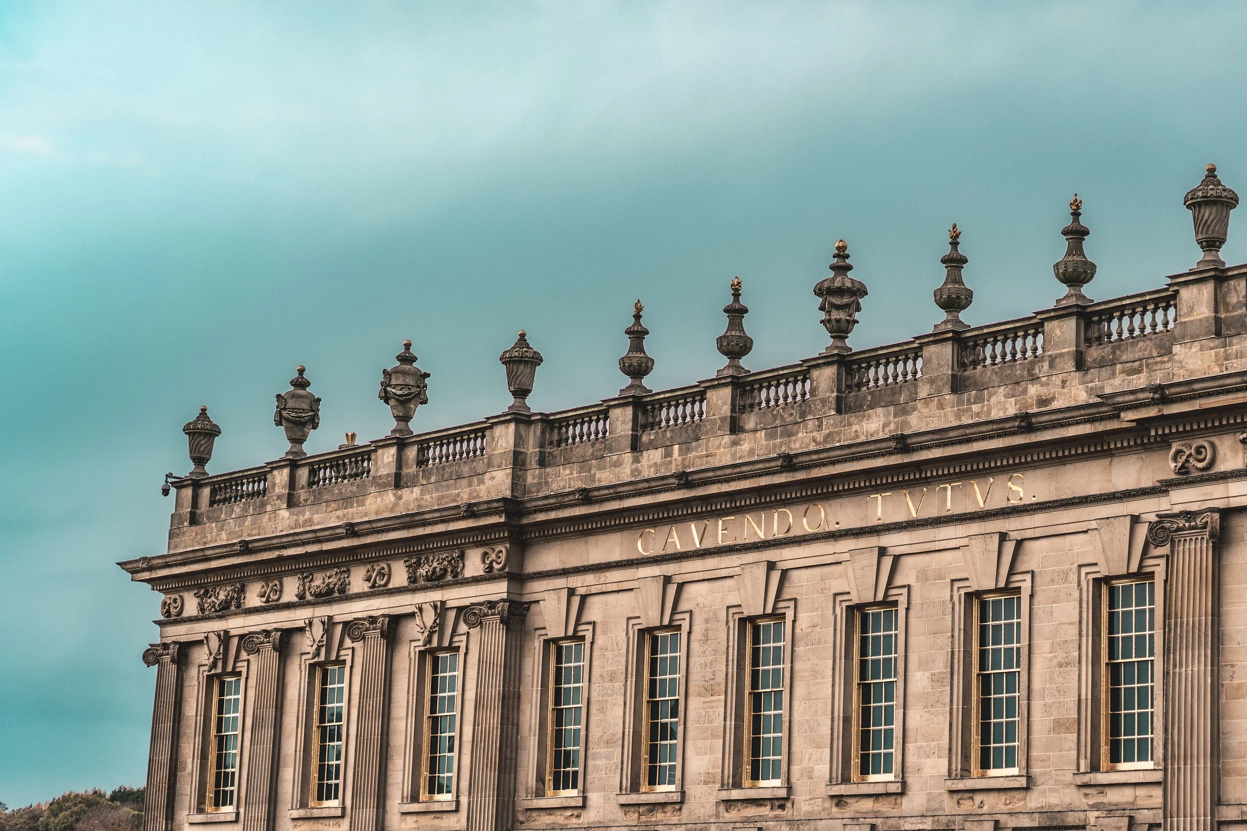 Close-up of a historic stone building with tall, narrow windows and ornate decorations, with Latin inscription 'CAVENDO TUTUS' on the facade, topped with decorative urns against a cloudy sky.
