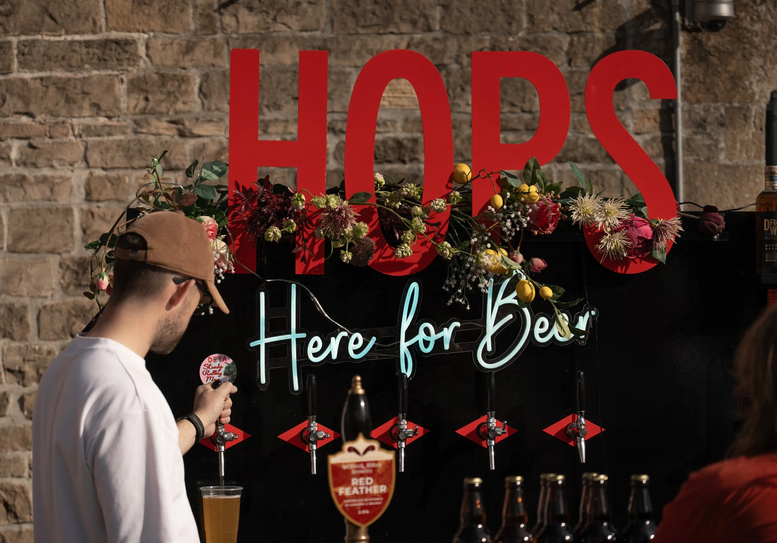 Bar with large red letters spelling 'HOPS' and a neon sign saying 'Here for Beer.' Several taps and bottles are visible, and a man is pouring beer into a plastic cup at a rustic brick wall background.
