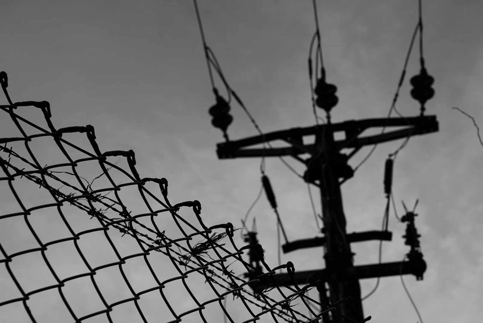 Silhouette of a utility pole and power lines behind a damaged chain-link fence against a cloudy sky.