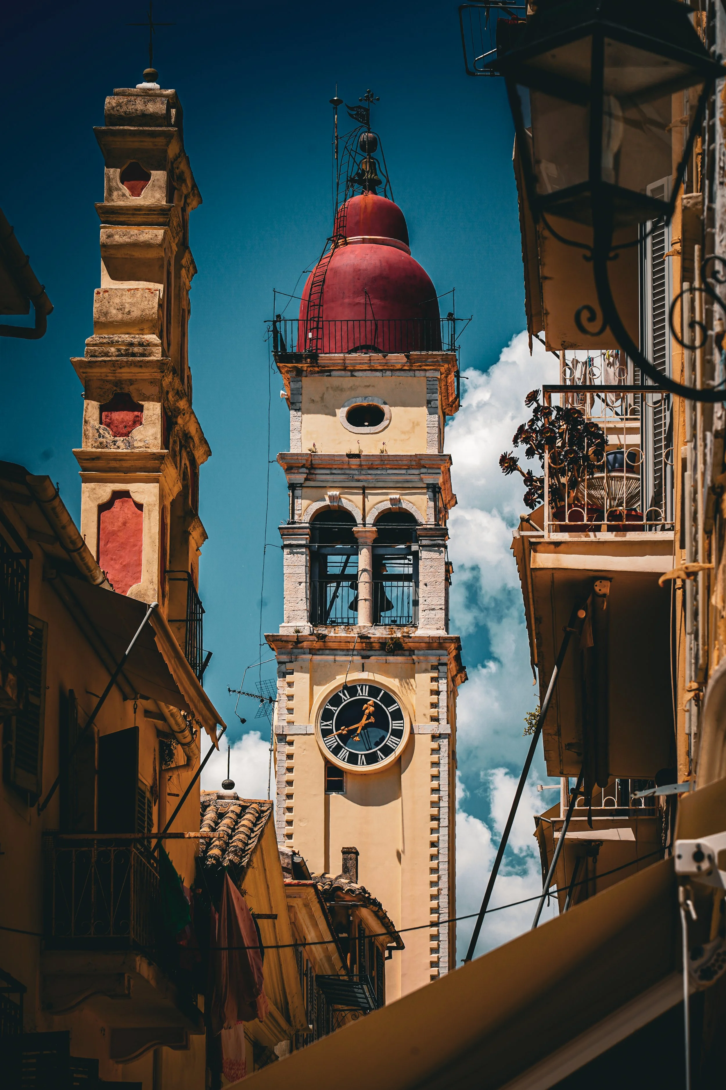 A historic clock tower with a red domed top, surrounded by buildings with balconies, under a blue sky with some clouds.