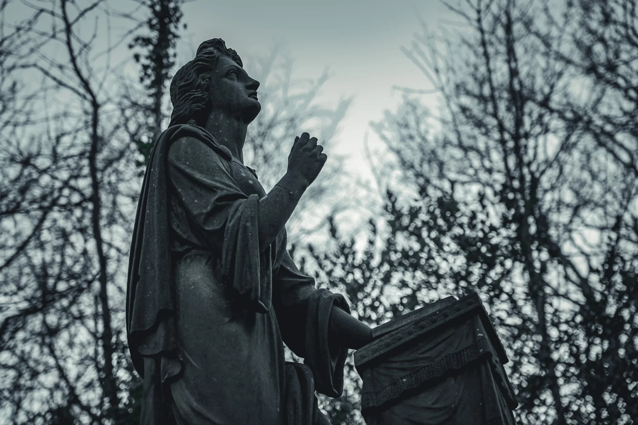 A black stone statue of a praying woman with her hands clasped together, set against a cloudy sky and leafless trees.