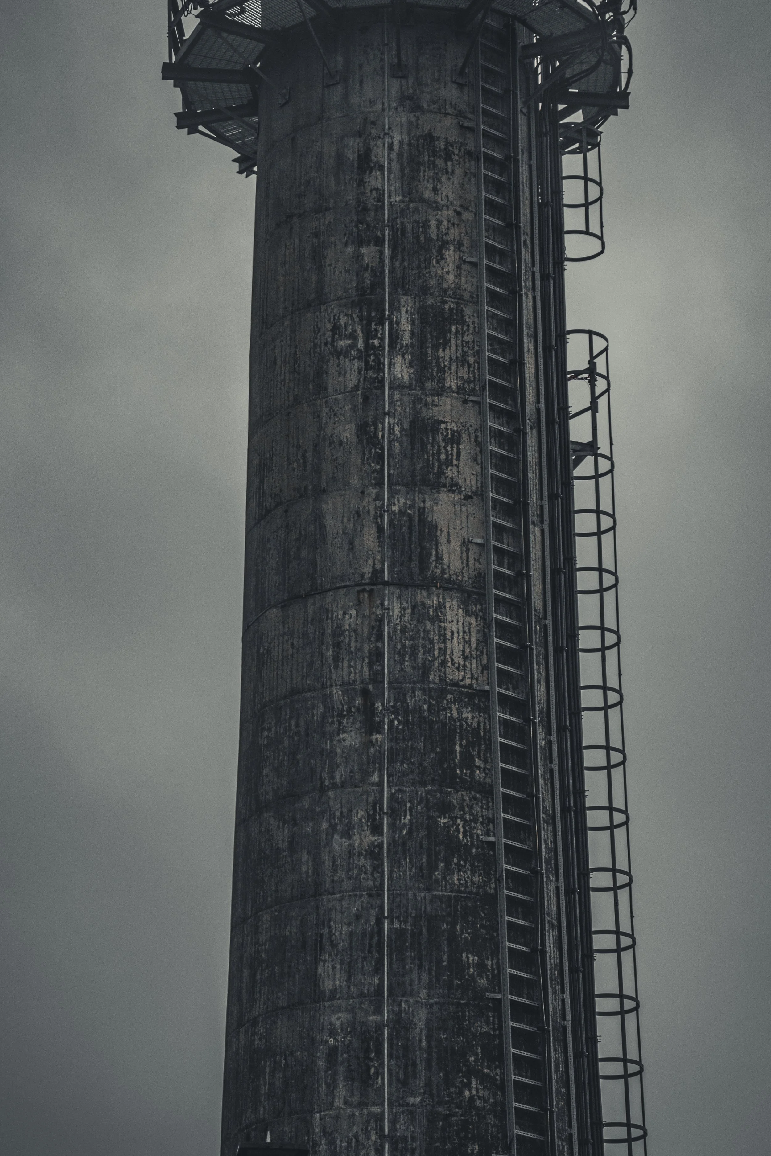 A tall, weathered industrial chimney with metal ladders and safety rings, set against a cloudy sky.