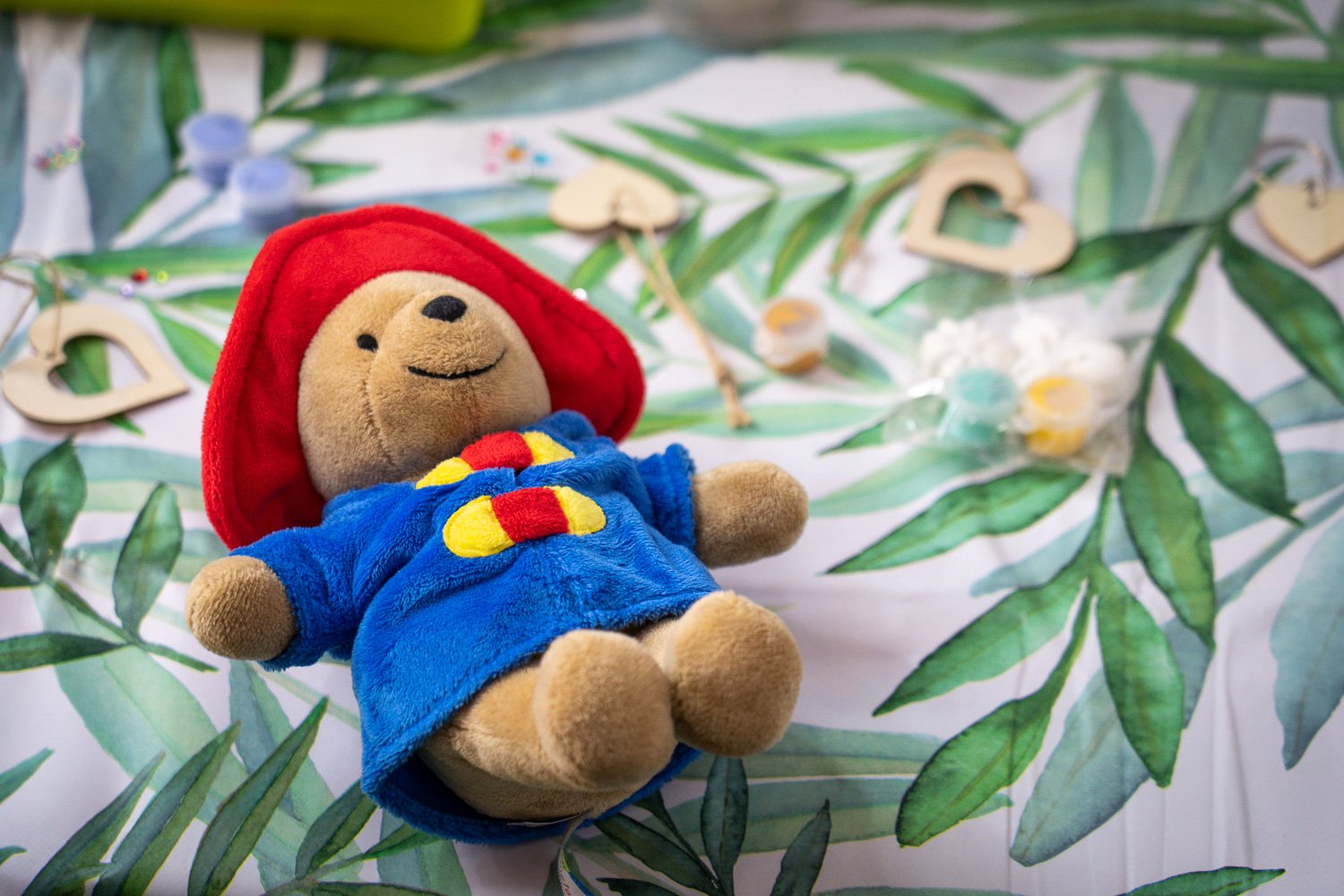 A stuffed teddy bear dressed in a blue and red outfit with a red hat, lying on a leafy patterned surface, surrounded by small heart-shaped cutouts and packages of gummies.