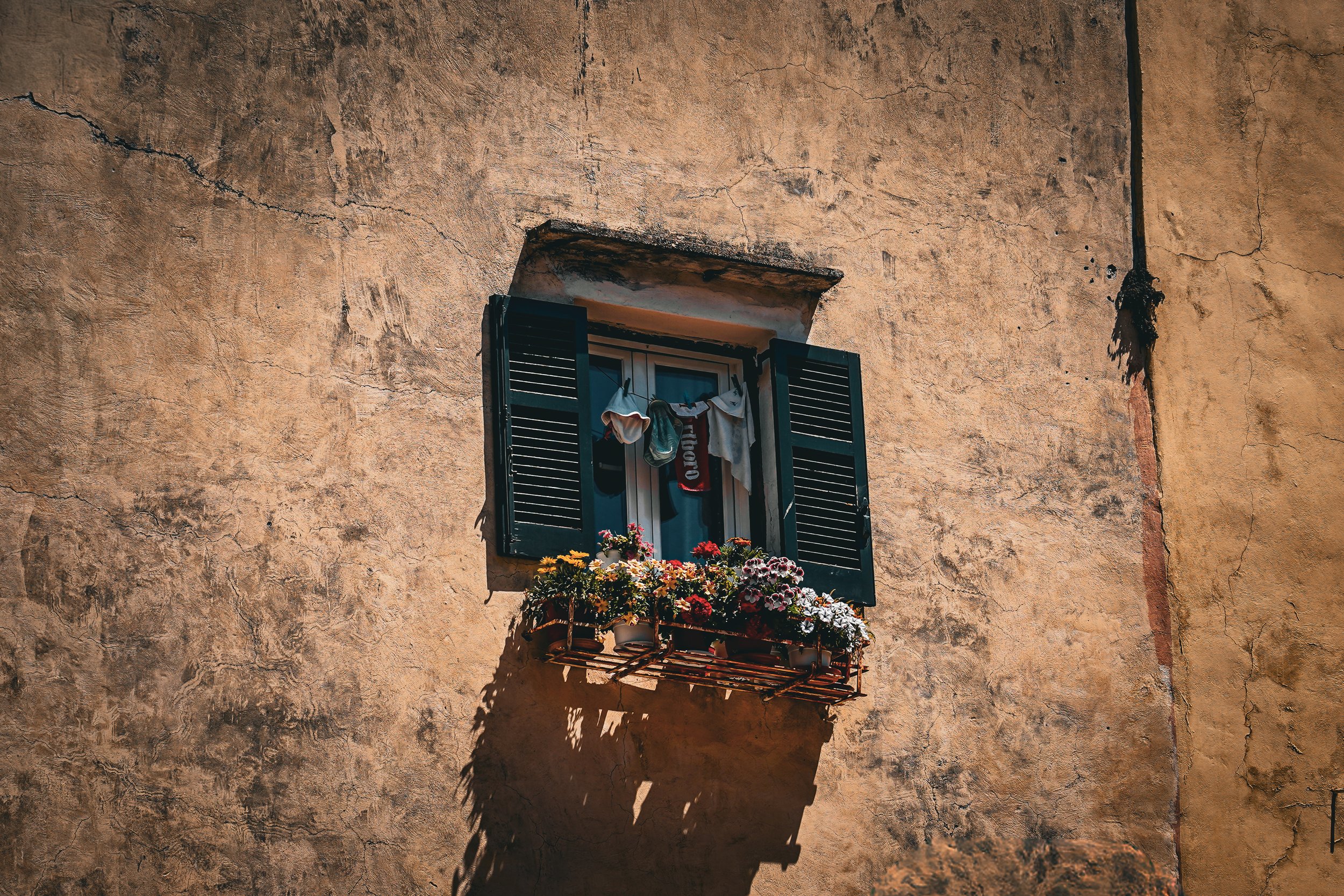 A window with black open shutters and laundry hanging on a white line, with a flower box filled with colorful flowers below on a textured, weathered wall.