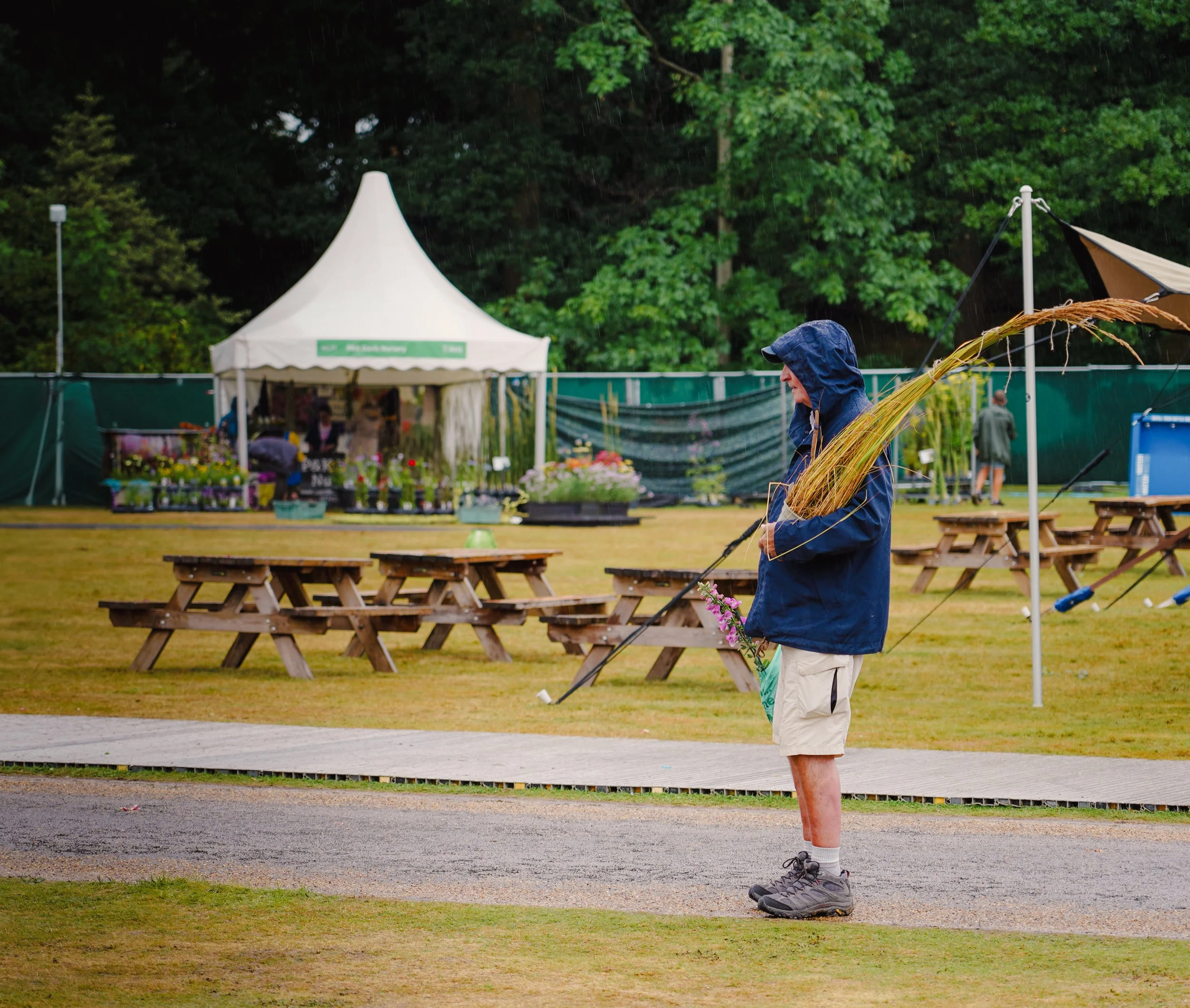 A man standing on a wet sidewalk carrying a gardening rake and a potted plant, in a park or festival setting with picnic tables, tents, and trees in the background during rainy weather.