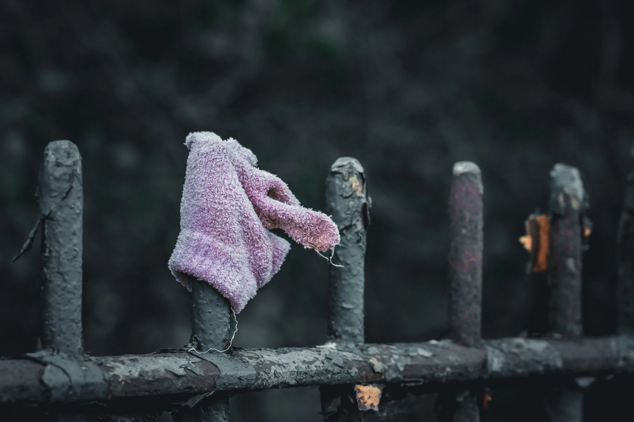 A small, pink stuffed animal wrapped in a fuzzy towel perched on a rusted, deteriorating metal fence in a dark outdoor setting.
