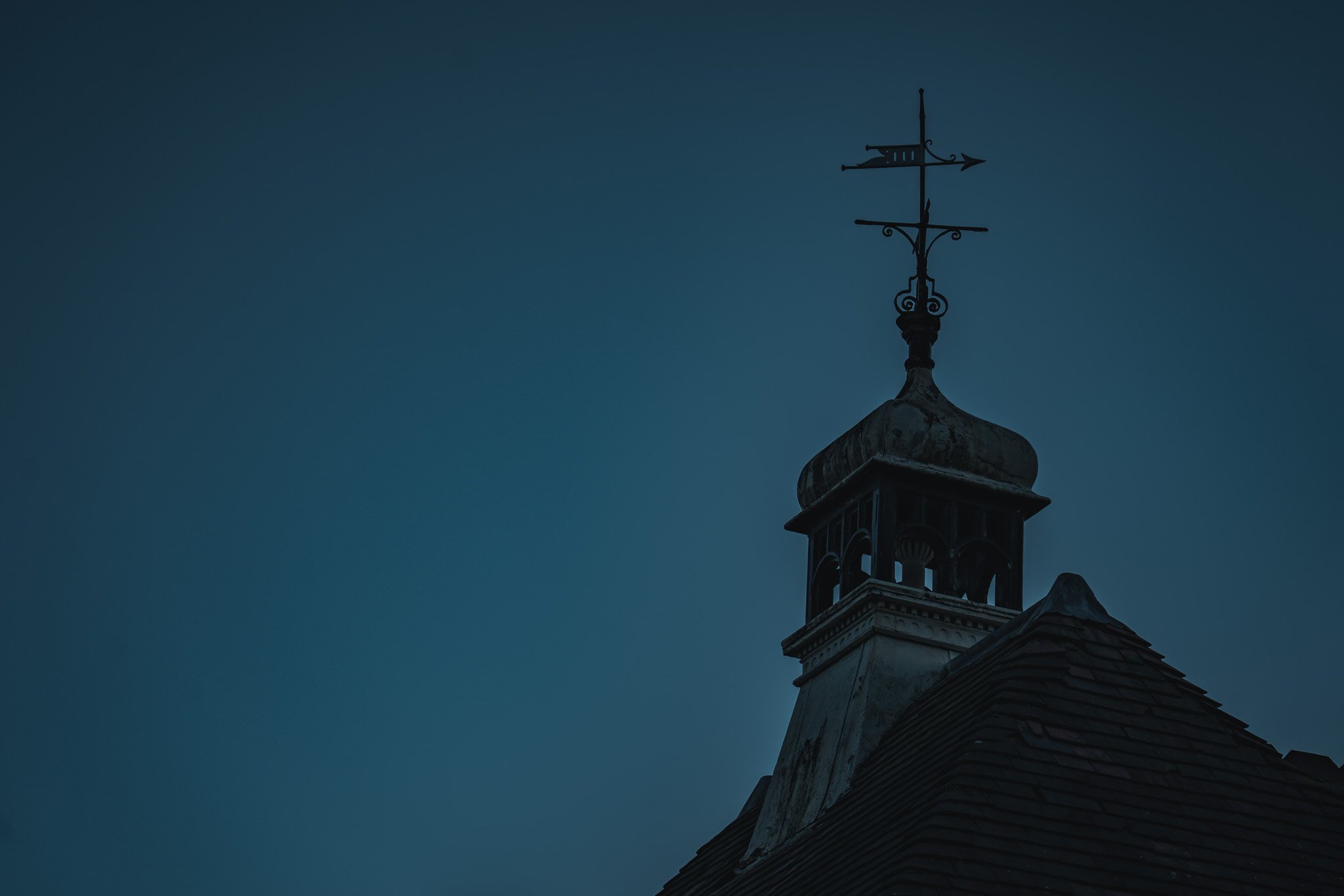 A church steeple with a weather vane on top against a darkening sky.