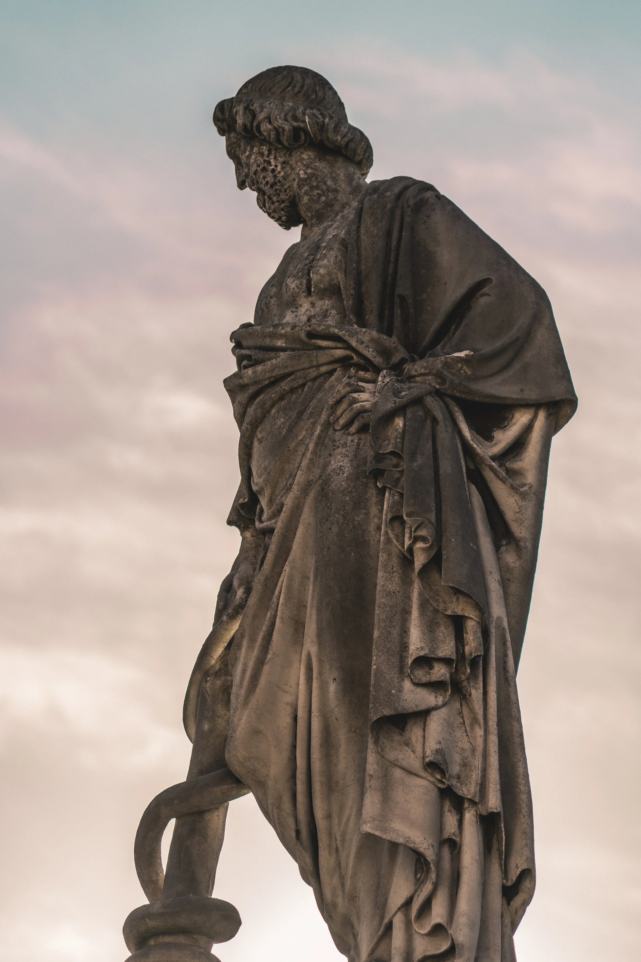A weathered stone statue of a bearded man looking down, with a draped robe, standing against a cloudy sky.