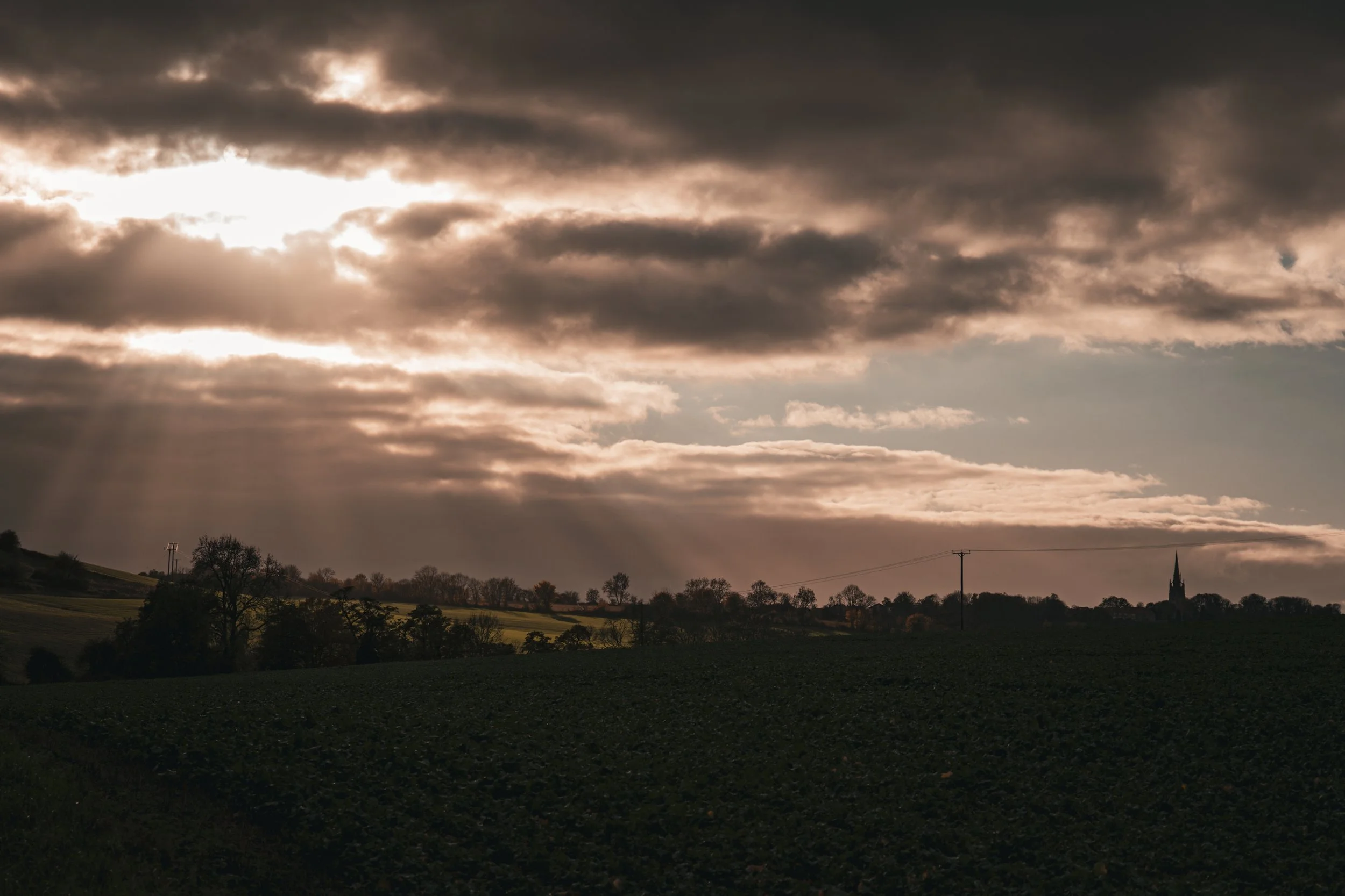 A rural landscape under a cloudy sky with rays of sunlight filtering through, showing fields, trees, telephone poles, and a church steeple in the distance.