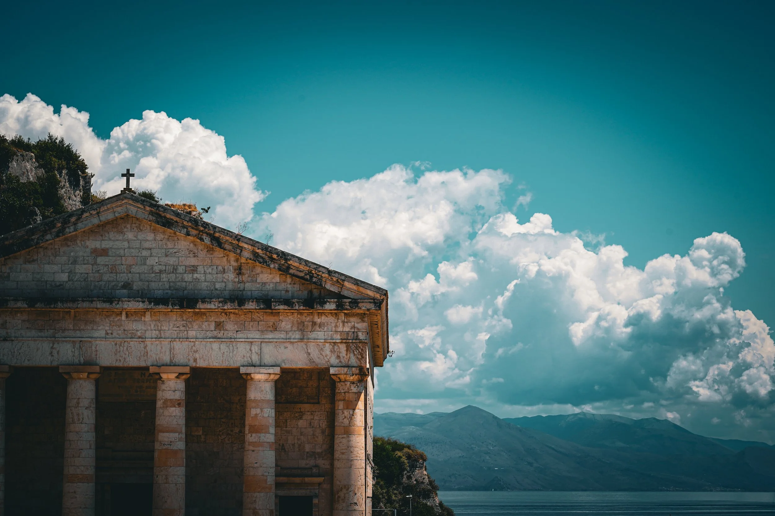 View of an old stone church with a cross on the roof, overlooking mountains and a body of water, under a sky with scattered clouds.