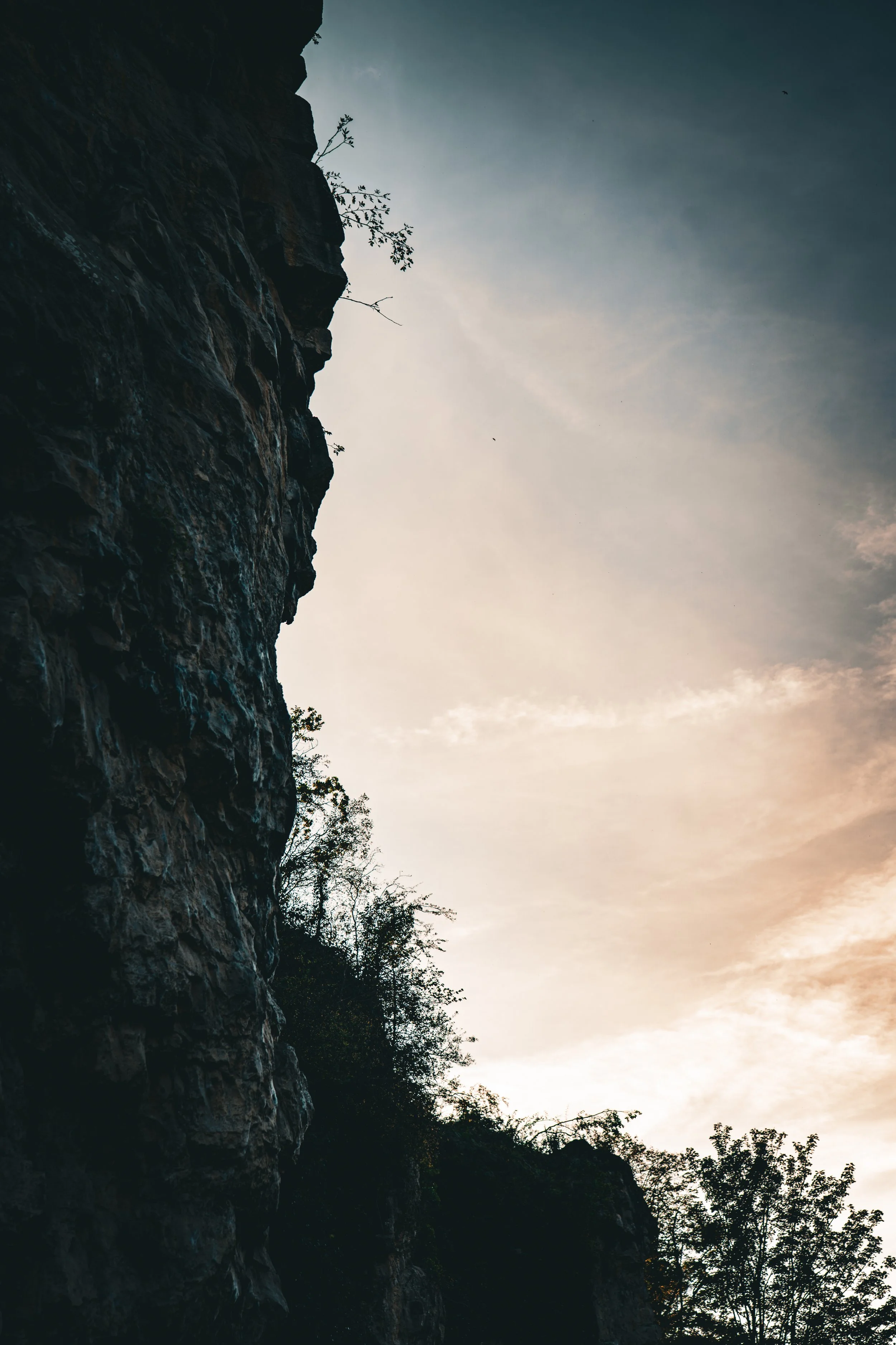 Silhouetted rocky cliff with sparse vegetation against a cloudy sky at sunset or sunrise.