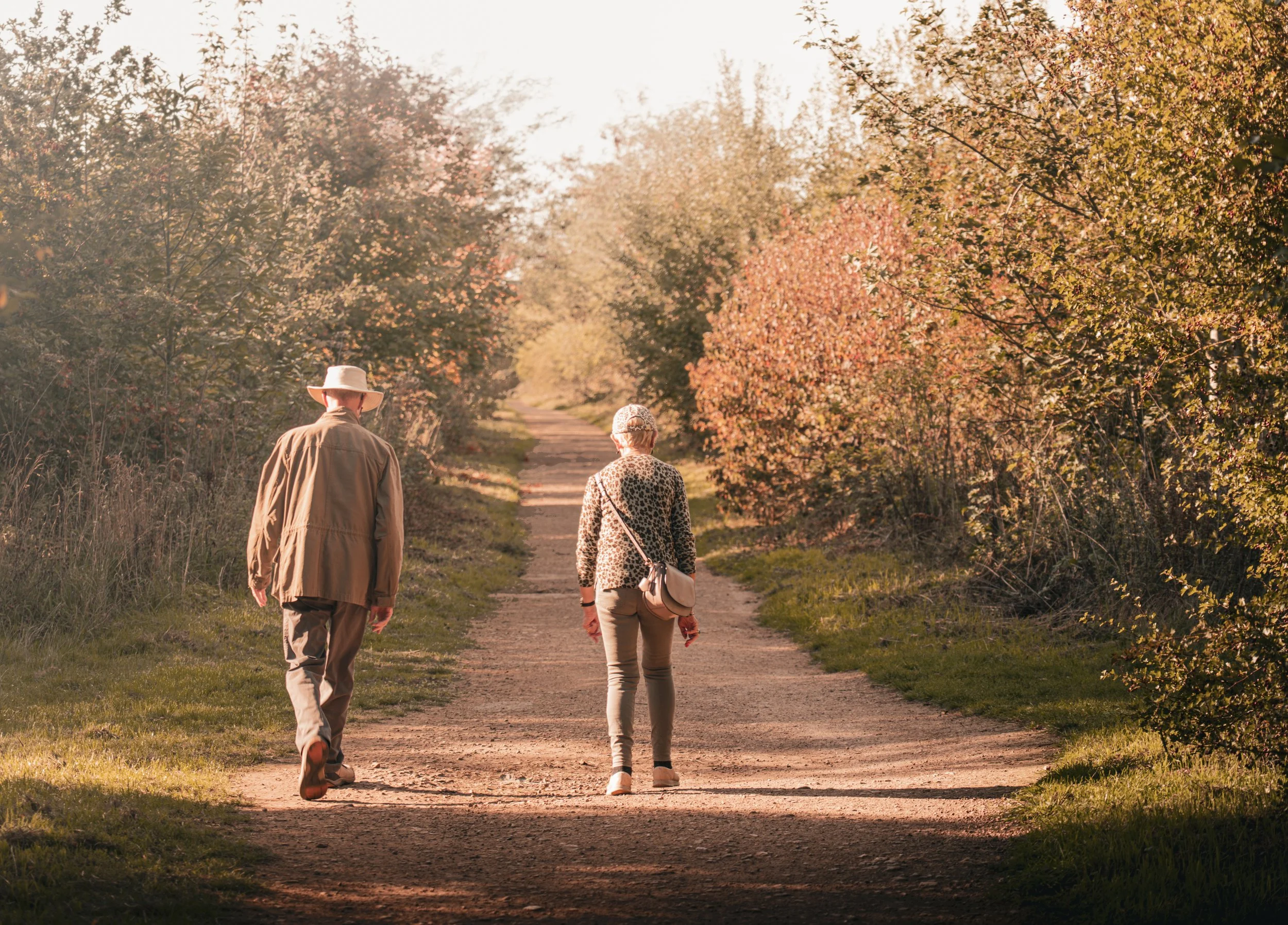Two people walking on a dirt path surrounded by trees and autumn foliage.