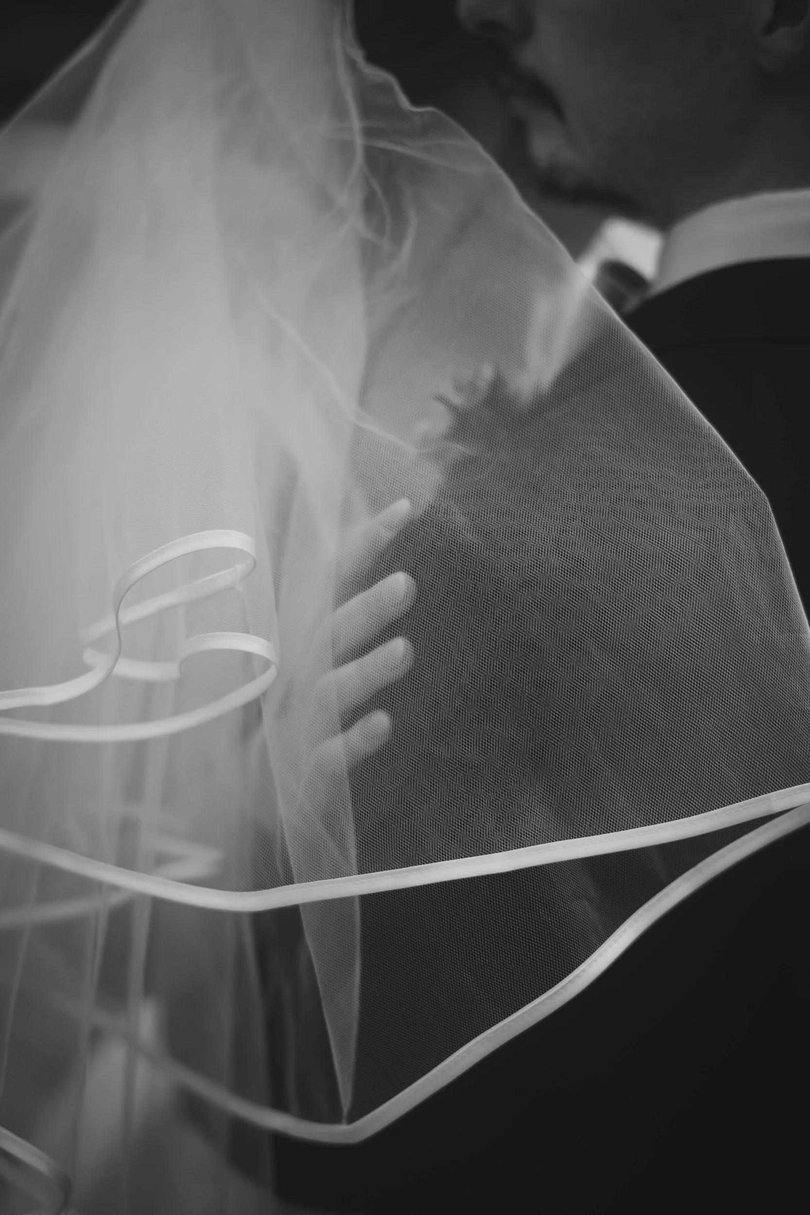 A woman wearing a wedding veil, seen gently touching her chest, with her face partially visible in the background, captured in black and white.