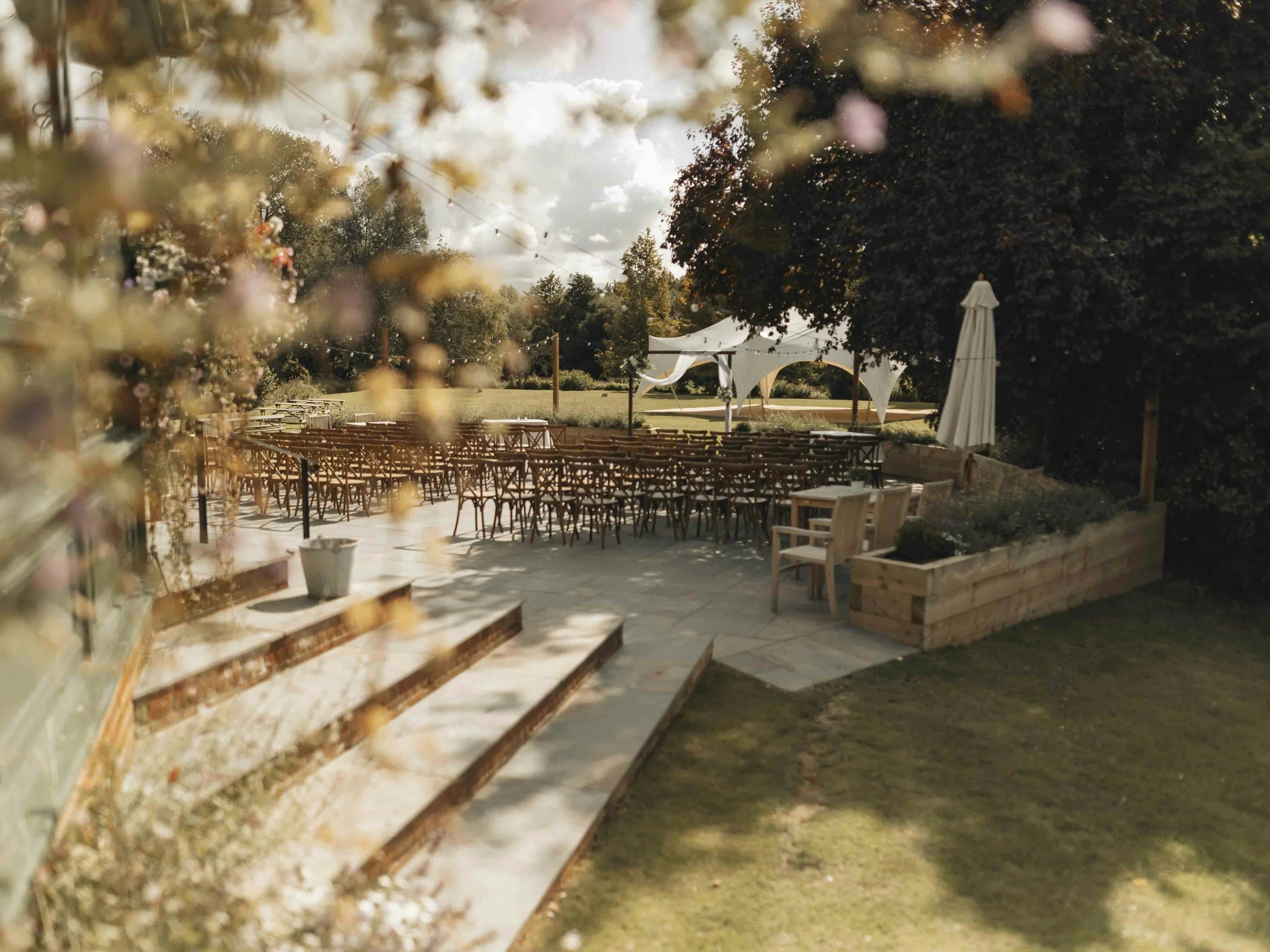Outdoor event space with folding chairs arranged on a patio, large white tents, and a surrounding garden with trees and flower beds.