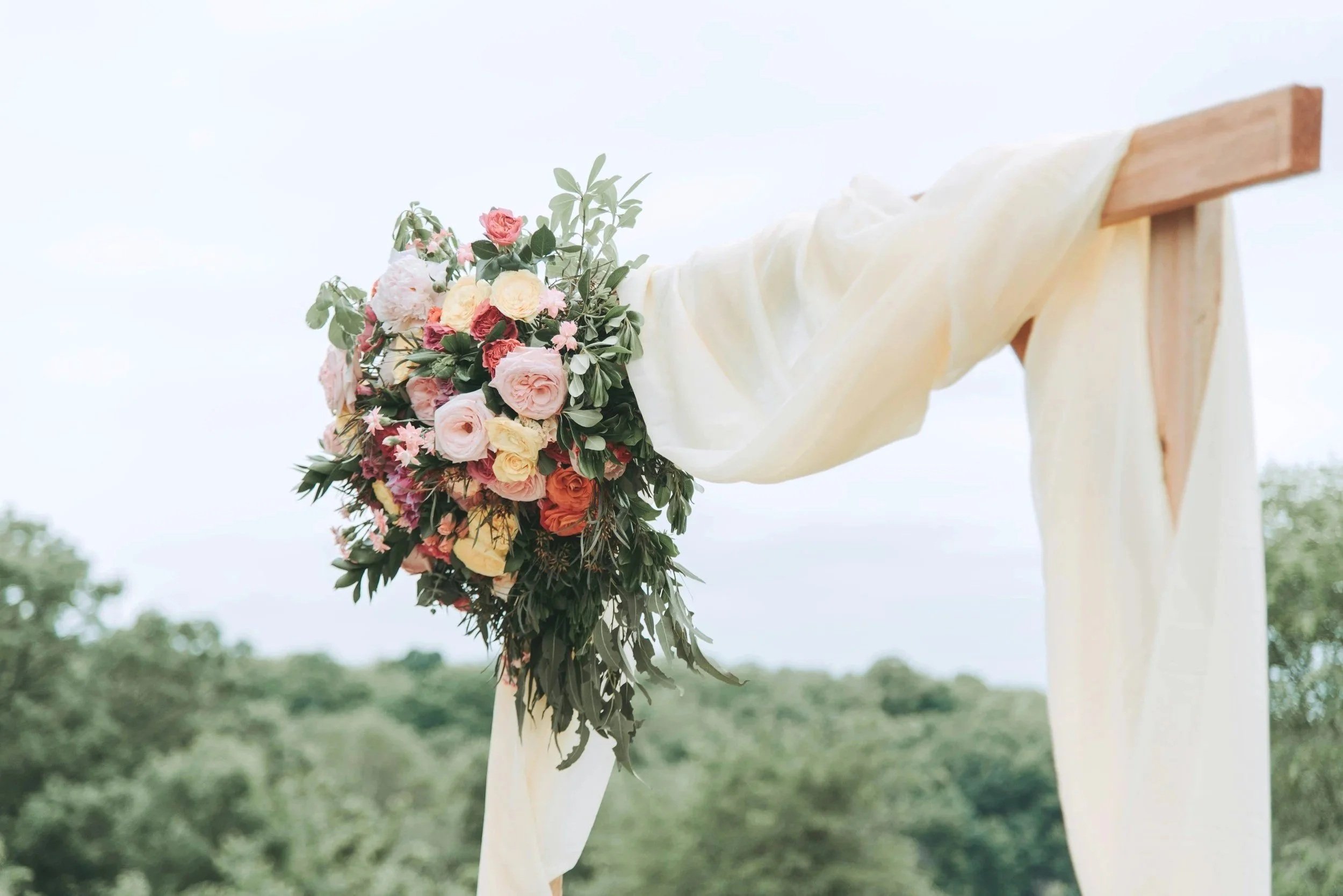 A floral wedding arch decorated with pink, yellow, and orange roses, draped with light cream fabric, set outdoors with trees in the background.
