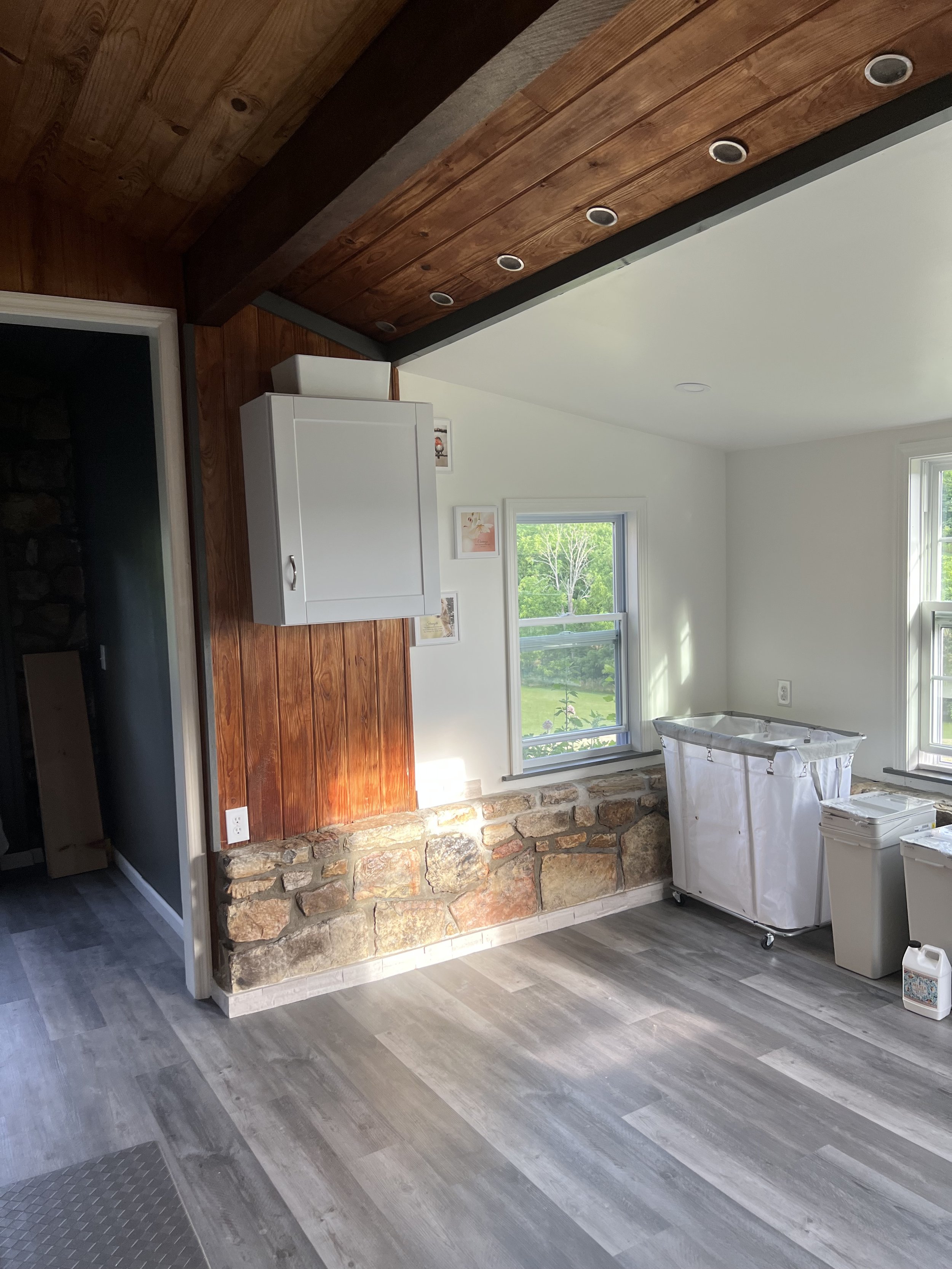 A room with white walls, large windows showing greenery outside, partially finished with a stone and wood wall, some painting supplies, and a laundry cart.