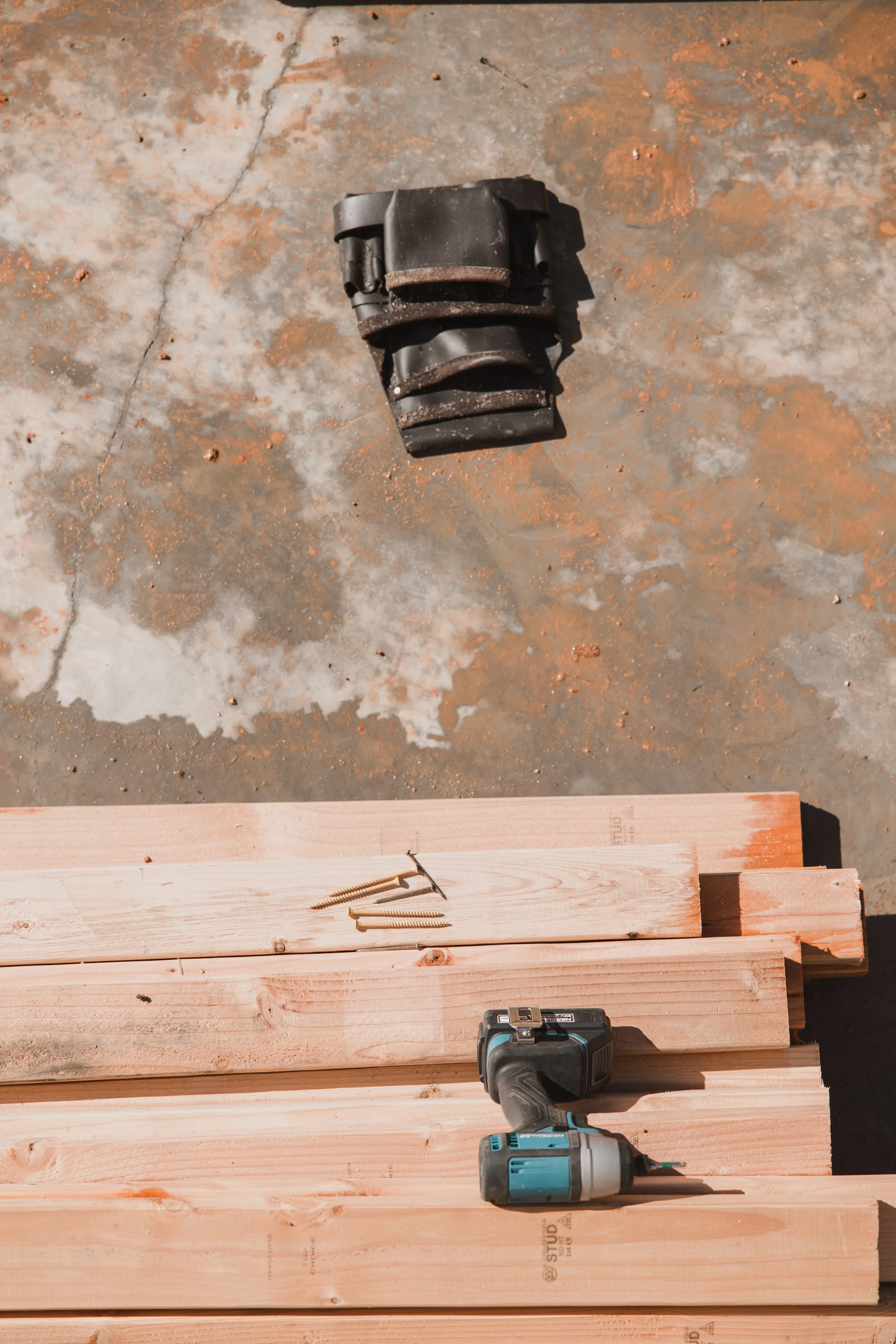 Construction tools including a cordless drill, a measuring tape, several wooden planks, screws, and clamps on a concrete floor.