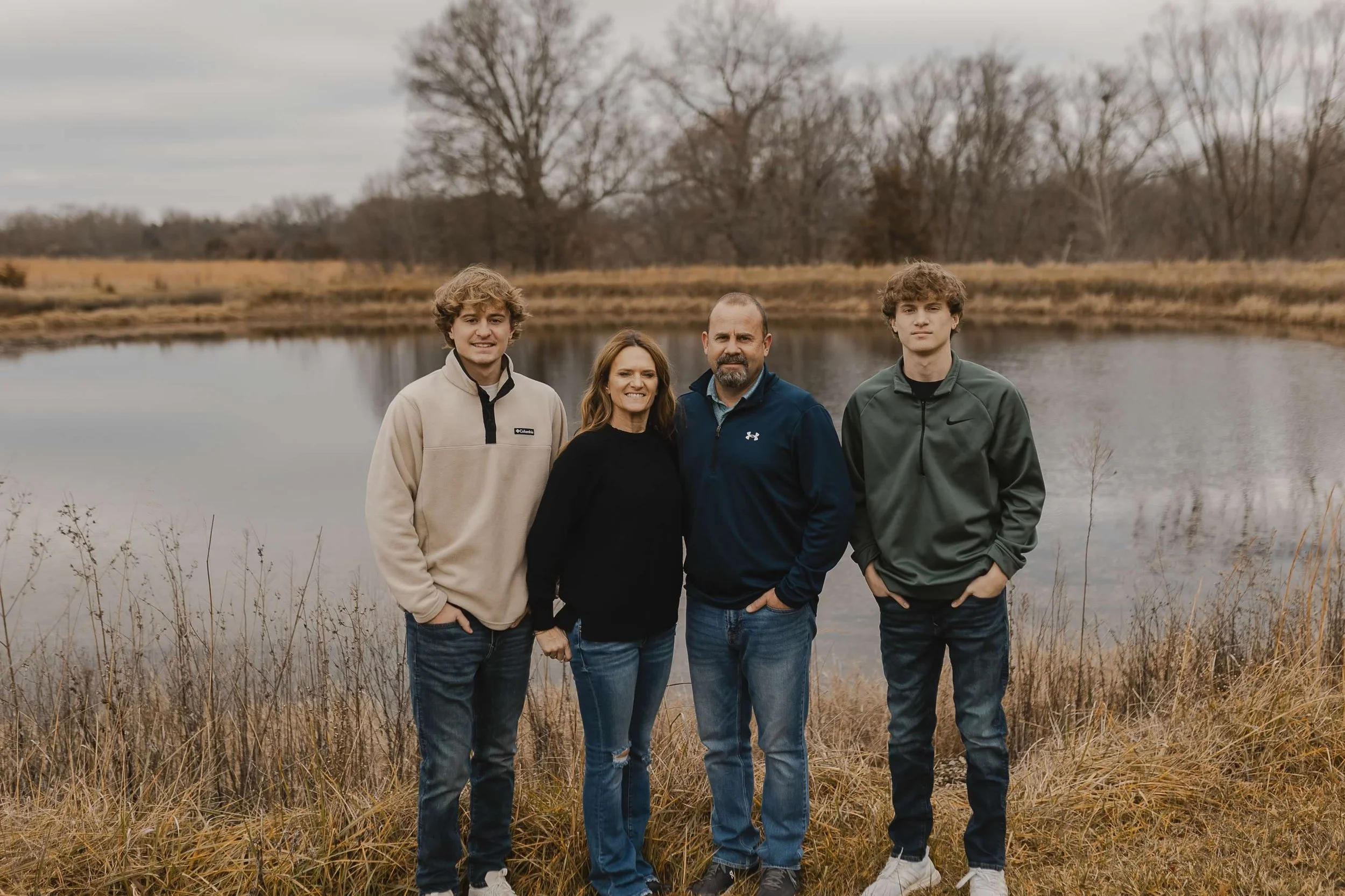Family of four standing outdoors near a pond during fall, with leafless trees and a cloudy sky in the background.