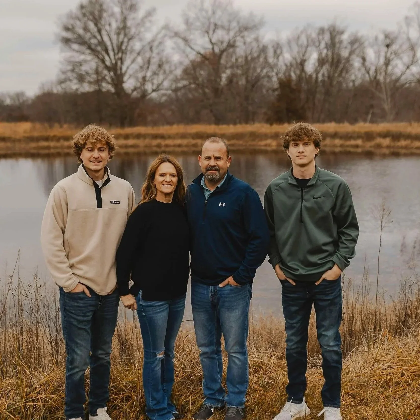 Family of four standing outdoors near a lake with trees in the background during autumn.