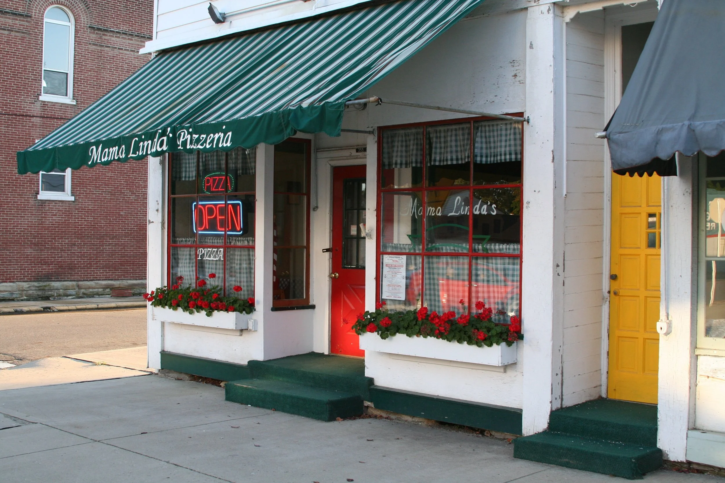 Front of a small pizzeria with a green awning reading 'Mama Linda's Pizzeria' and neon signs, surrounded by flower boxes with red flowers.