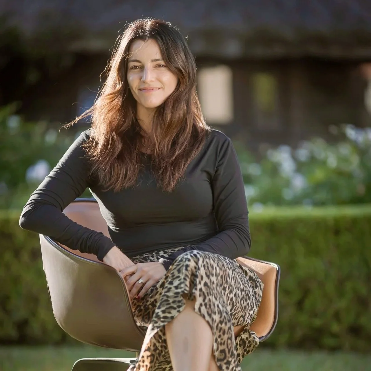 A woman with long brown hair seated outdoors on a modern chair, smiling with a lush green background. SONA Co-founder headshot