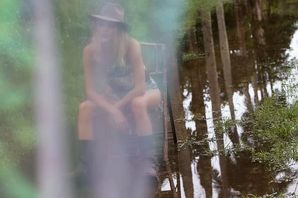A woman sitting outdoors near a body of water with tall trees reflected in the water, wearing a hat and boots.