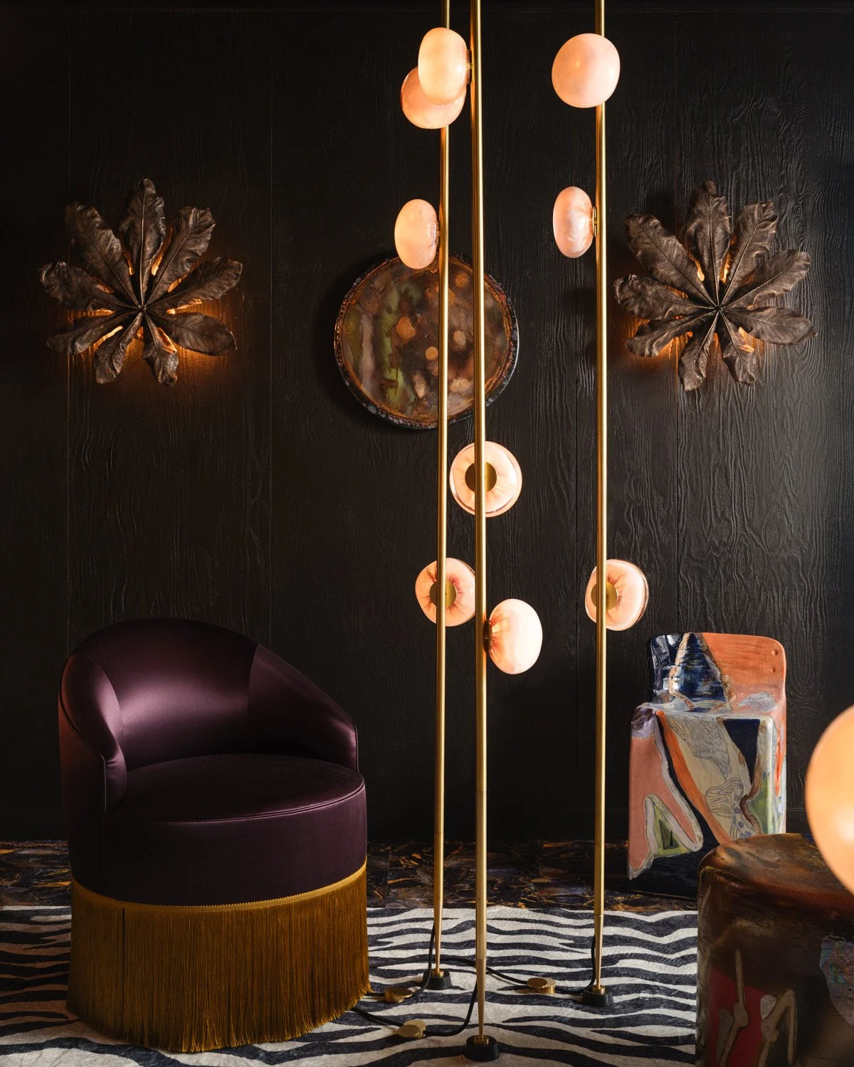 Interior decor with purple velvet chair with gold fringe, modern standing lamp with pink globes, textured black wall with bronze leaves, abstract painted side table, and striped rug.