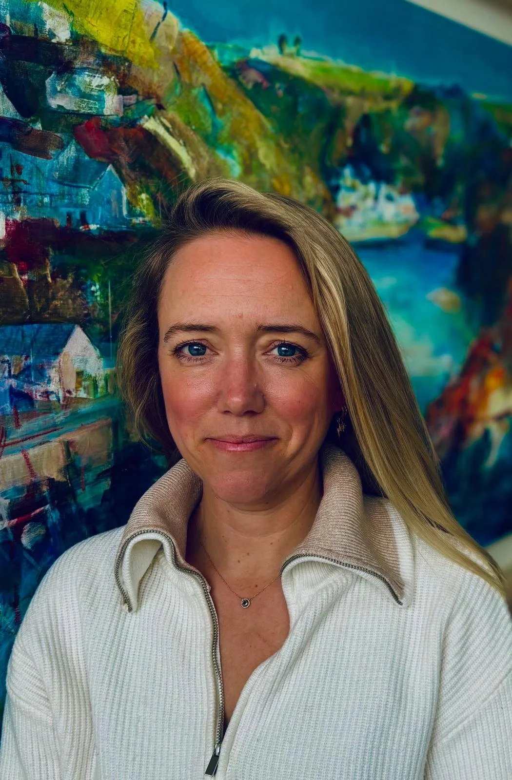 A woman with blonde hair and blue eyes, wearing a beige zip-up sweater and a necklace, standing in front of an abstract colorful painting.