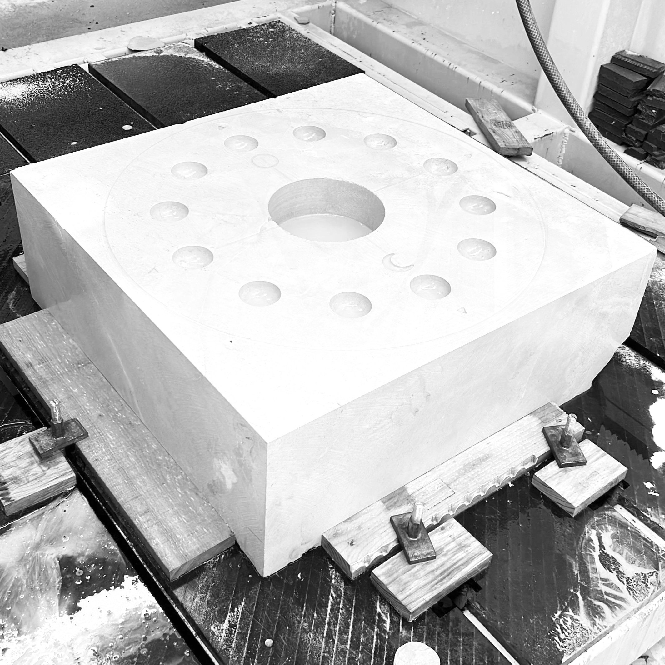 A large square block of marble or similar stone with circular holes drilled into the top surface, placed on a workbench surrounded by clamps and tools in a workshop.