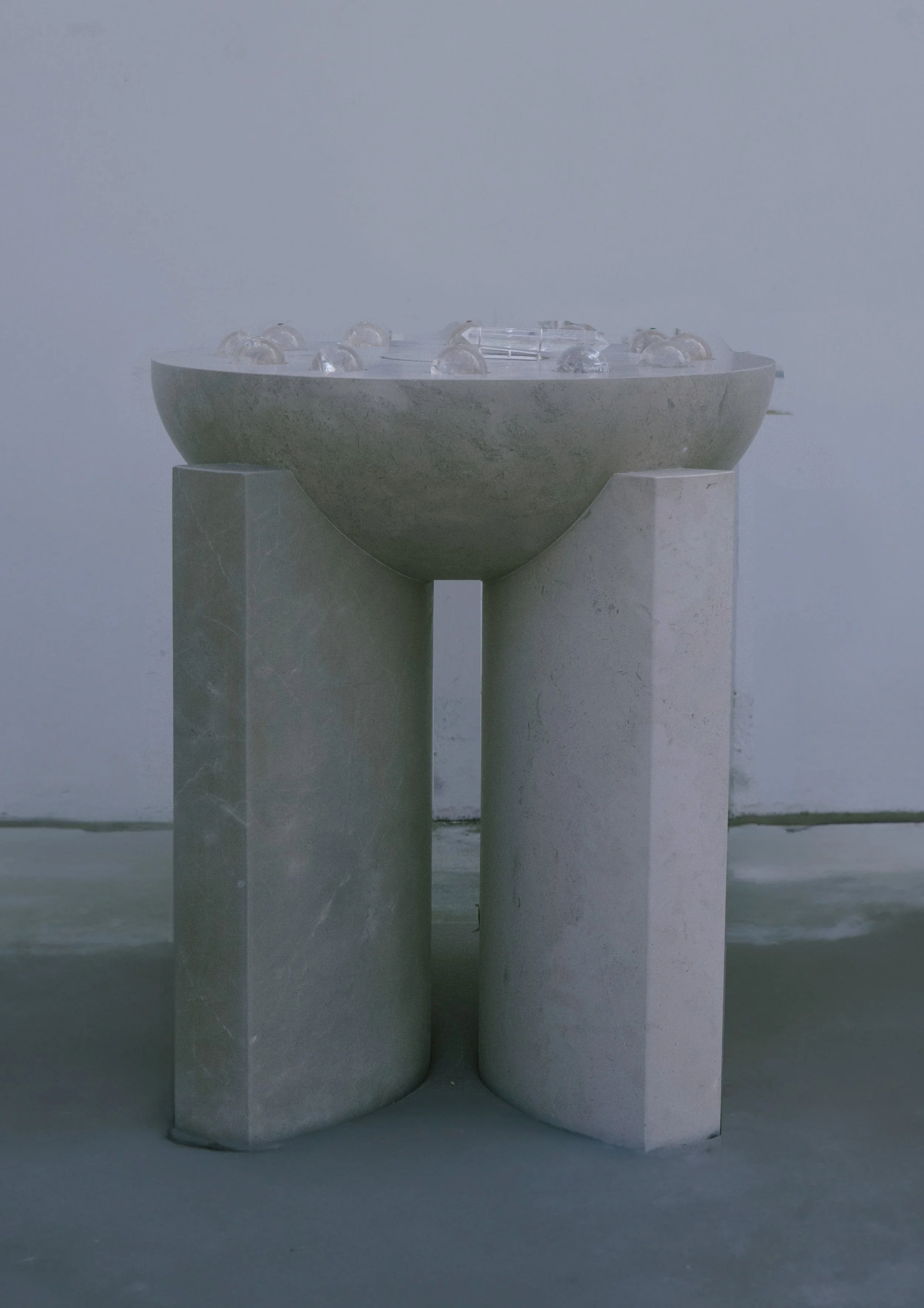 A minimalist sculpture or art installation featuring a round, shallow bowl resting on two vertical rectangular stone pedestals. The bowl contains several clear glass spheres or objects.