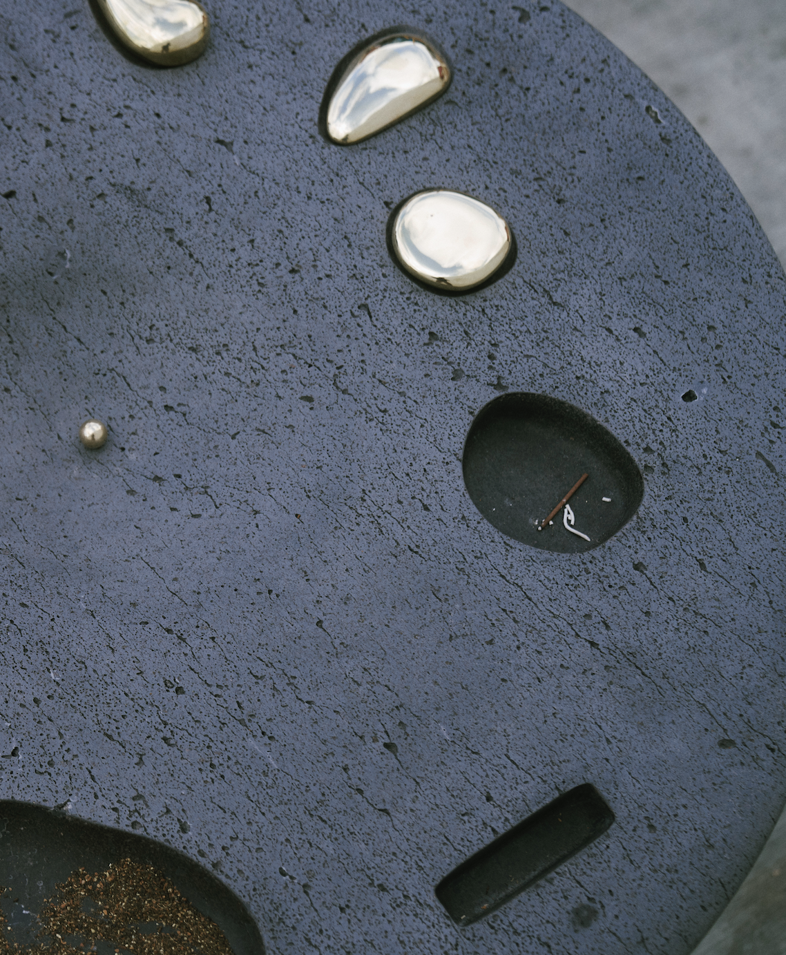 Close-up of a textured dark surface with reflective metallic drops of water, a small round metallic ball, and a circular indentation containing various debris.