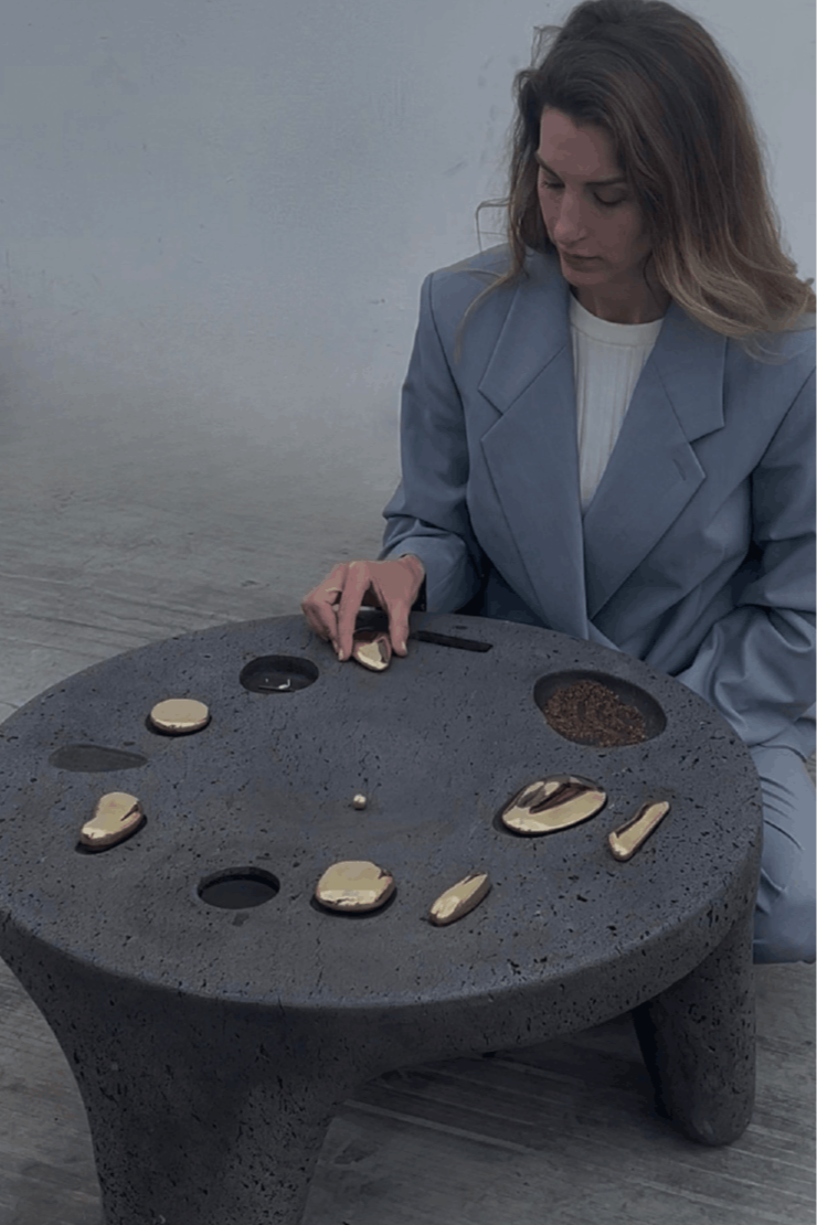 A woman in a light blue blazer sitting at a round stone table with various small objects and some black and brown substances on it.
