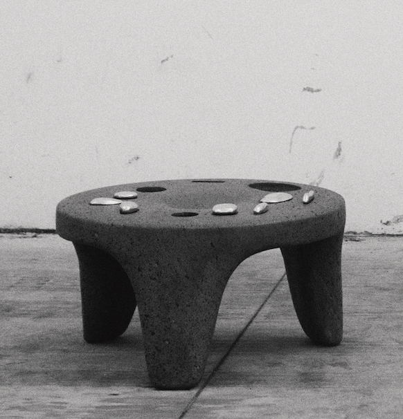 A small, three-legged stone stool with a round top, placed on a wooden floor against a white wall, with several small stones on its surface.