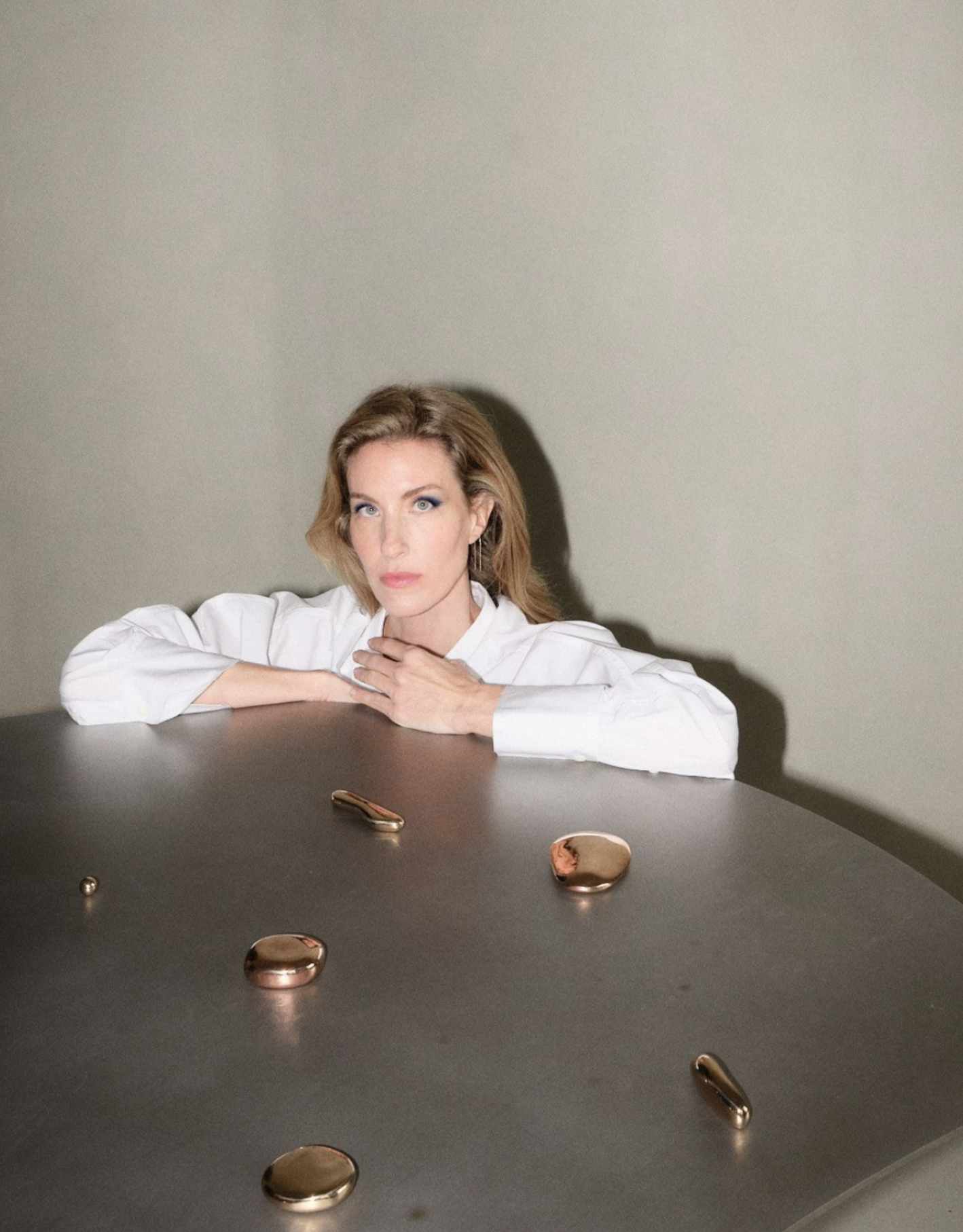 A woman with blonde hair resting her chin on her crossed arms on a dark table, looking directly at the camera. The table has several small, shiny, metallic objects scattered on it. The background is a plain, light-colored wall.