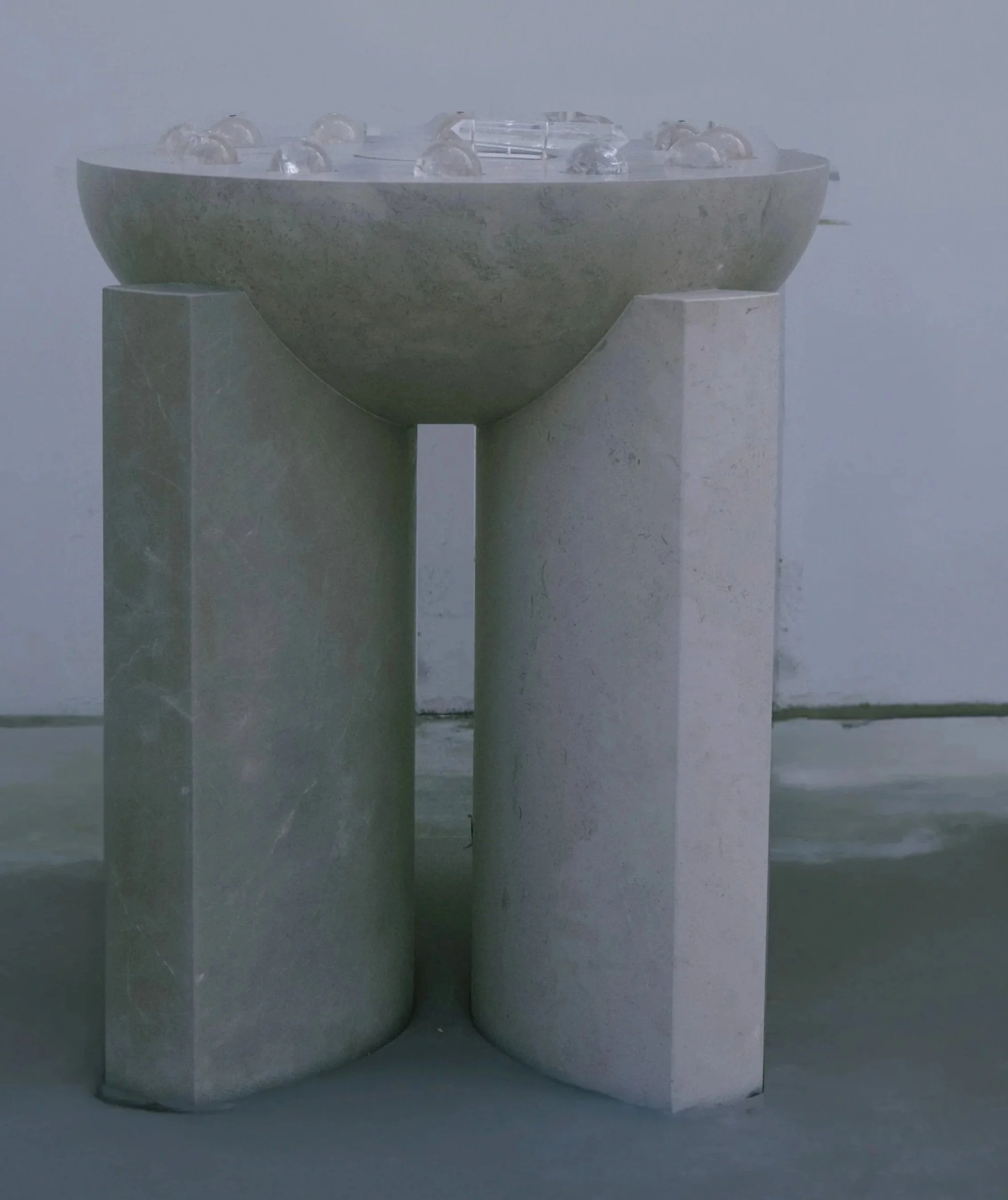 A modern sculpture composed of two large, rectangular stone blocks supporting a semi-spherical stone bowl on top, with small glass or crystal objects inside the bowl.
