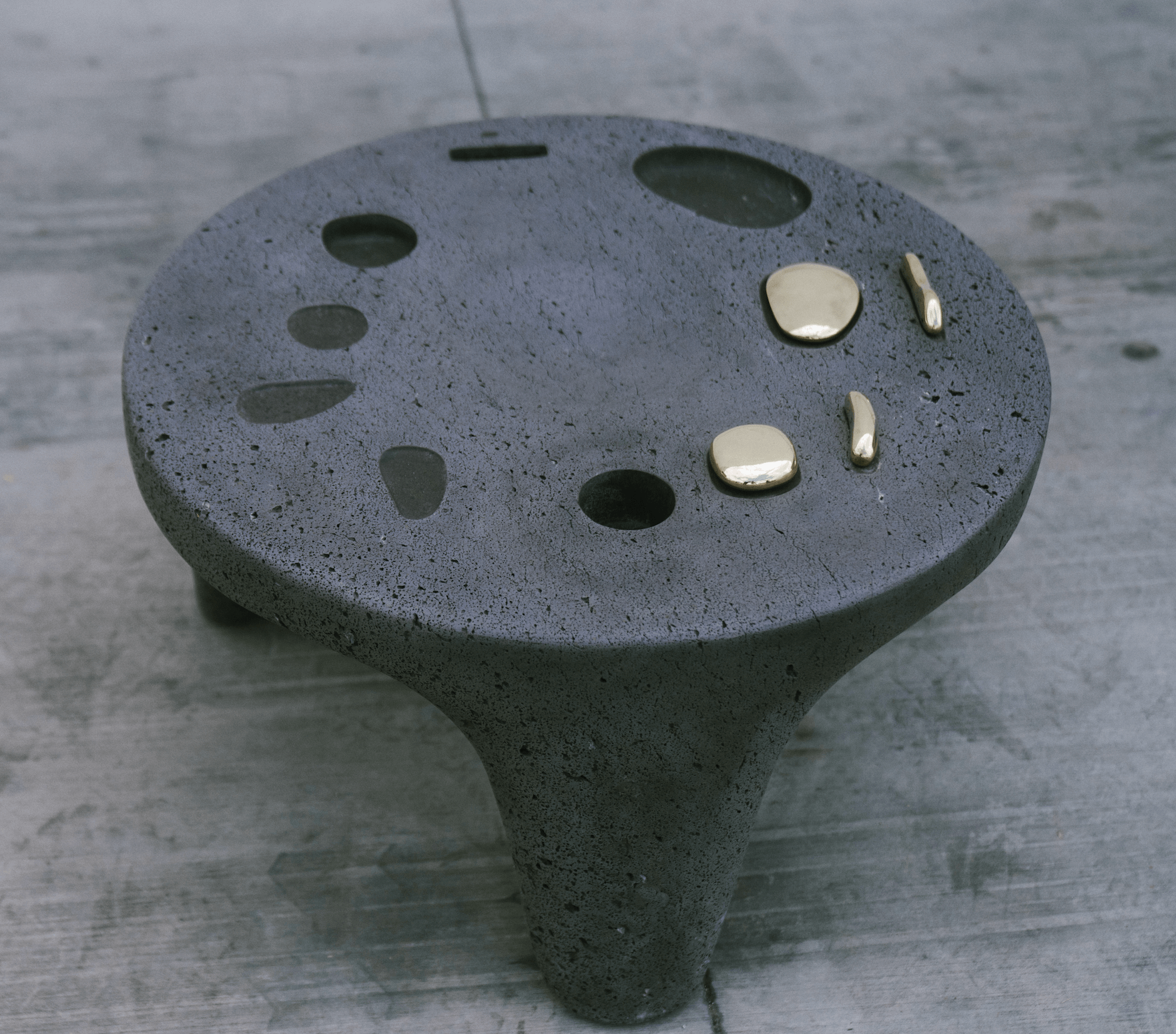 A black, textured, stool-shaped object with metallic accents and cut-out shapes on its surface, placed on a wooden floor.