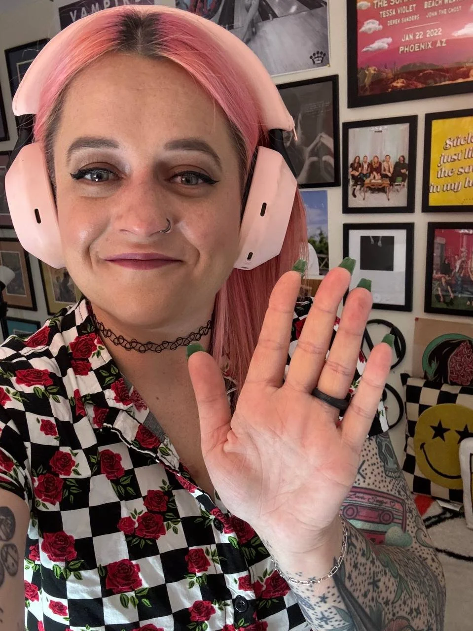 A woman with pink hair wearing large pink headphones, a black choker, and a checkered shirt with roses, smiling and waving. She has tattoos on her arm and a nose ring. The background features framed pictures, posters, and colorful pillows.
