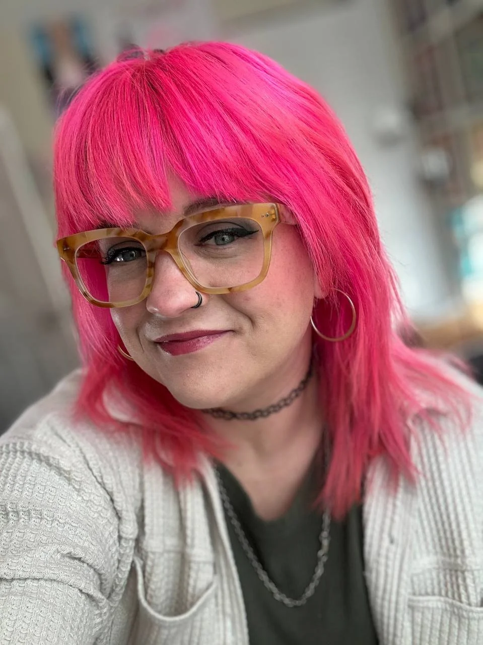 A woman with bright pink hair, wearing glasses with a tortoiseshell frame, a nose ring, hoop earrings, a black choker, and a necklace, smiling at the camera.