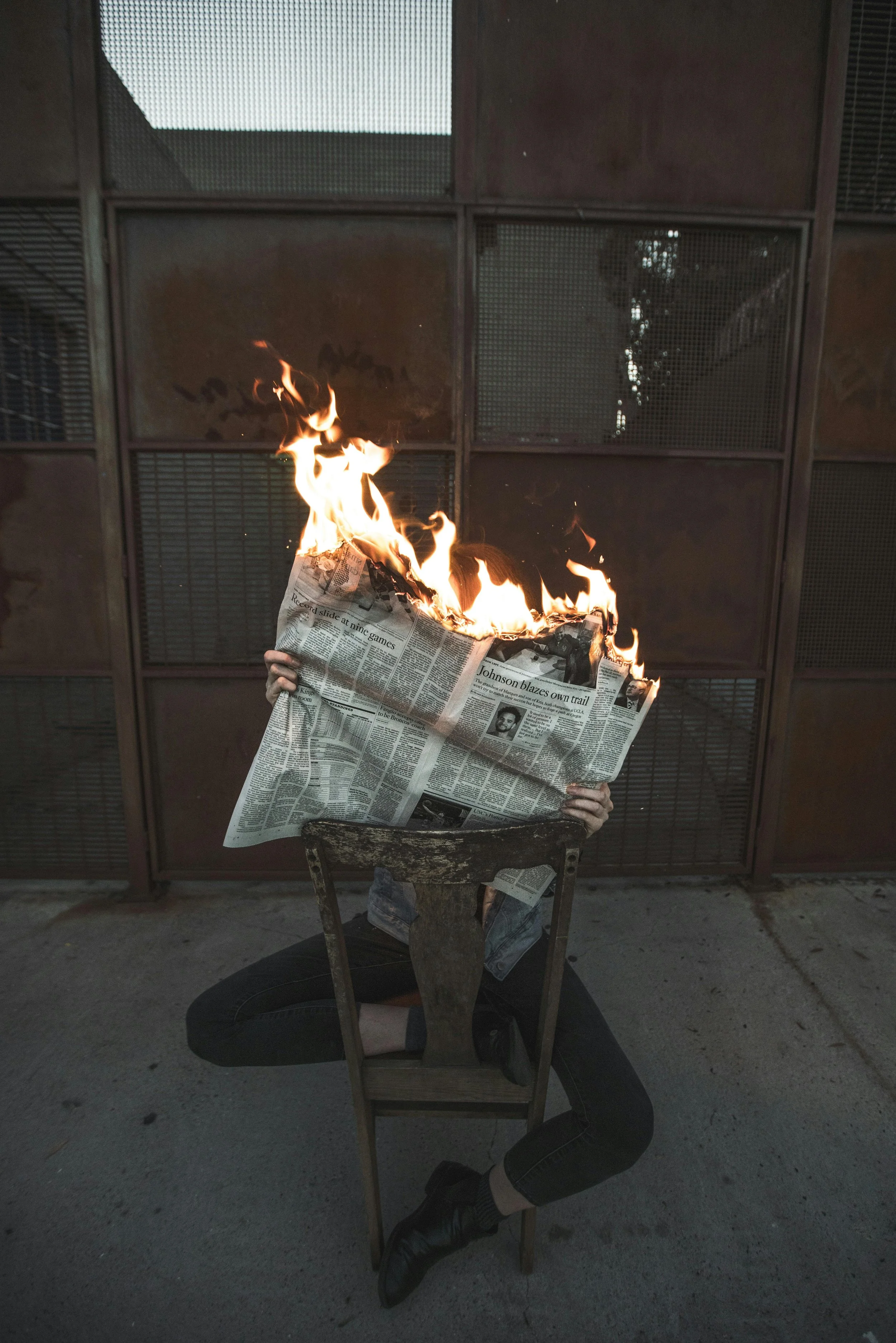 A person of an unknown gender is sitting backwards on a wooden chair and is holding up a burning newspaper which is covering their face.