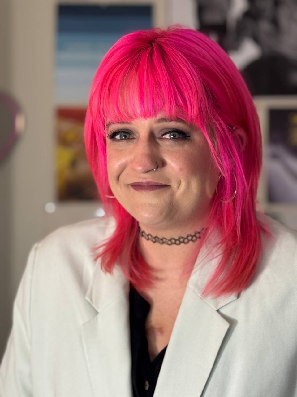 Close-up portrait of a woman with bright pink hair, wearing a white blazer, black shirt, choker necklace, and hoop earrings, indoors with blurred background.