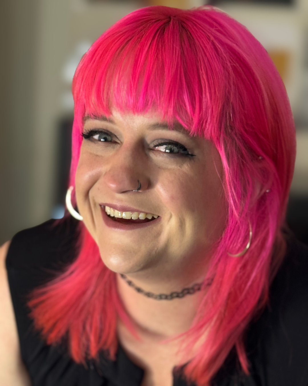 A smiling woman with bright pink hair, wearing hoop earrings, a black choker, and a black top, inside a room.