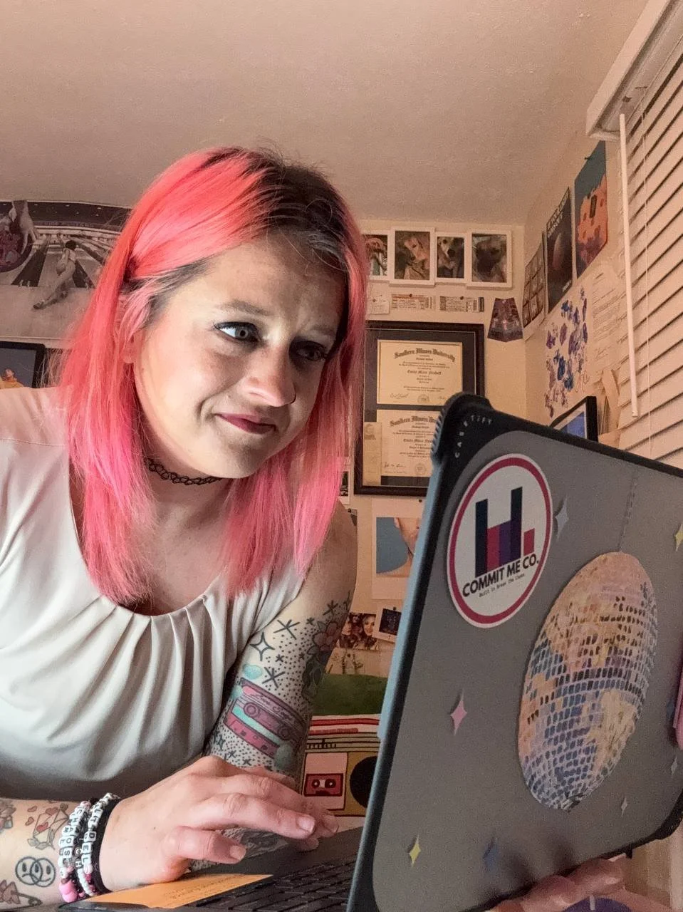 A woman with pink hair looking at her laptop, which has stickers, including a 'Commit Me Co.' logo. She has tattoos on her arm and is in a room decorated with framed certificates, posters, and photos.