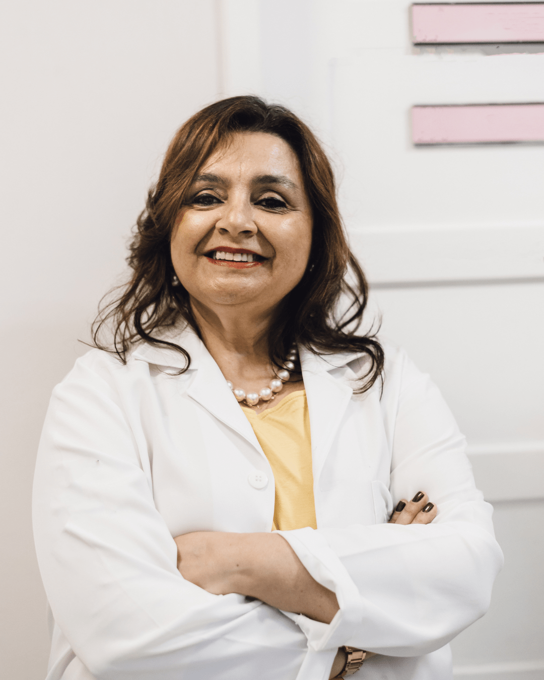A confident woman with shoulder-length brown hair, wearing a white lab coat, pearl necklace, and a yellow shirt, standing with arms crossed in a clinical setting.