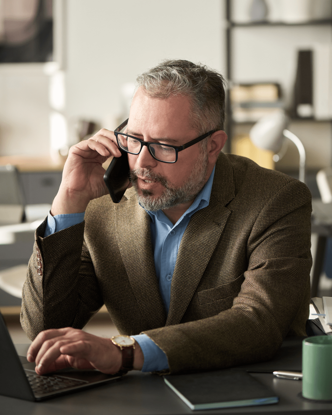 A headshot of a businessman on the phone, wearing black-rimmed glasses, and a brown suit with a blue shirt.