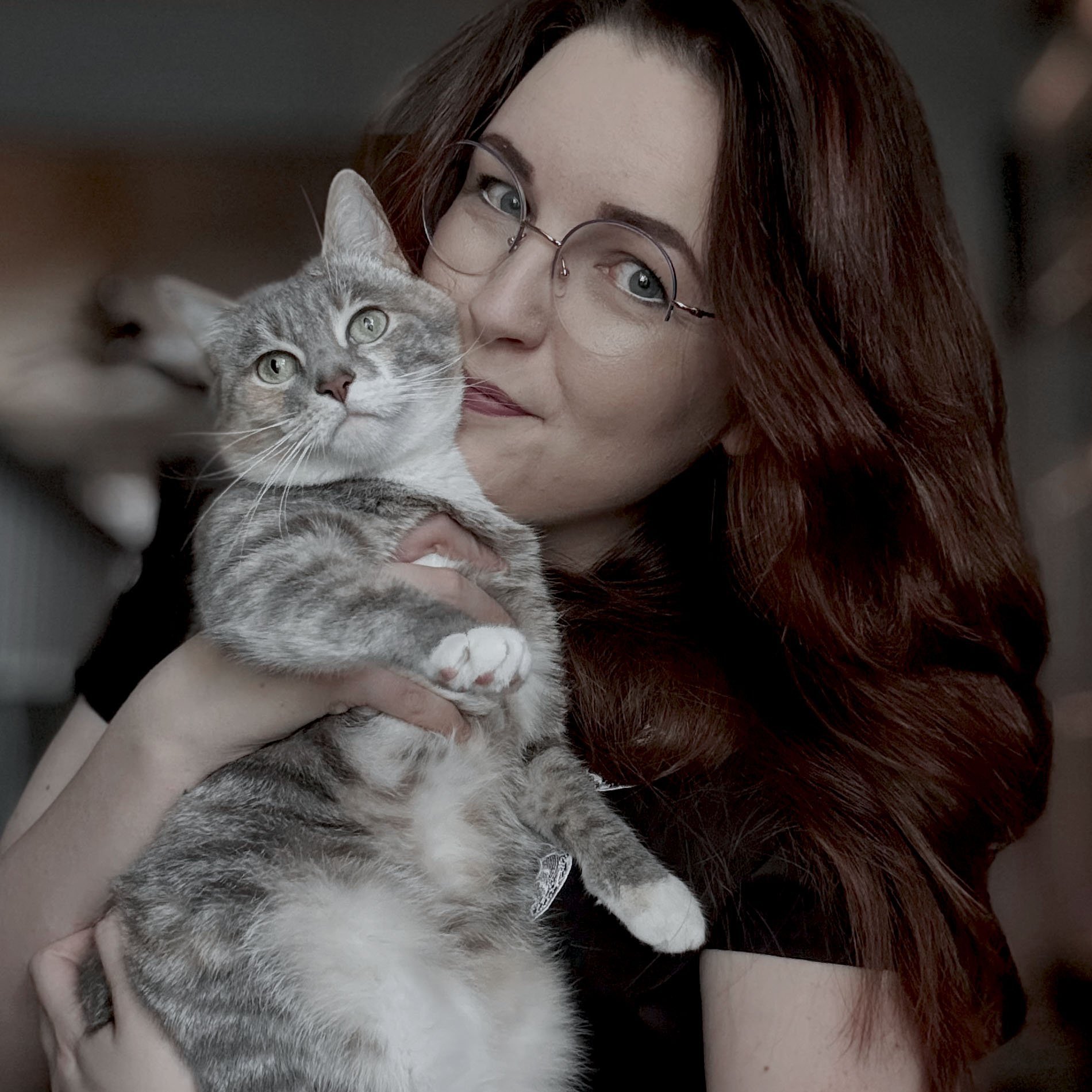 A woman with red hair and glasses is holding a gray and white tabby kitten close to her face, both looking into the camera.