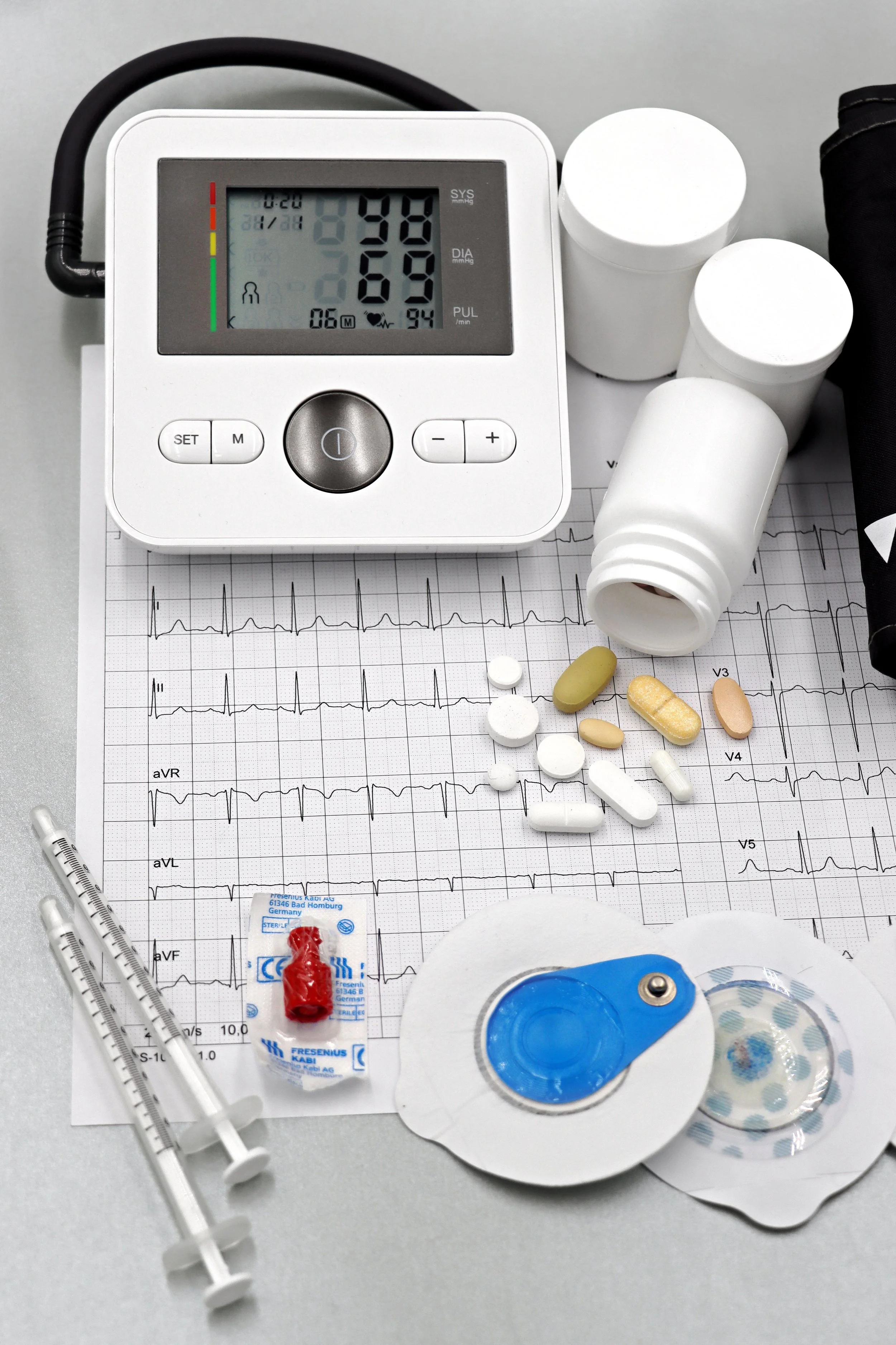Medical equipment including a digital blood pressure monitor, medications, an ECG printout, syringes, and a blood pressure cuff on a table.