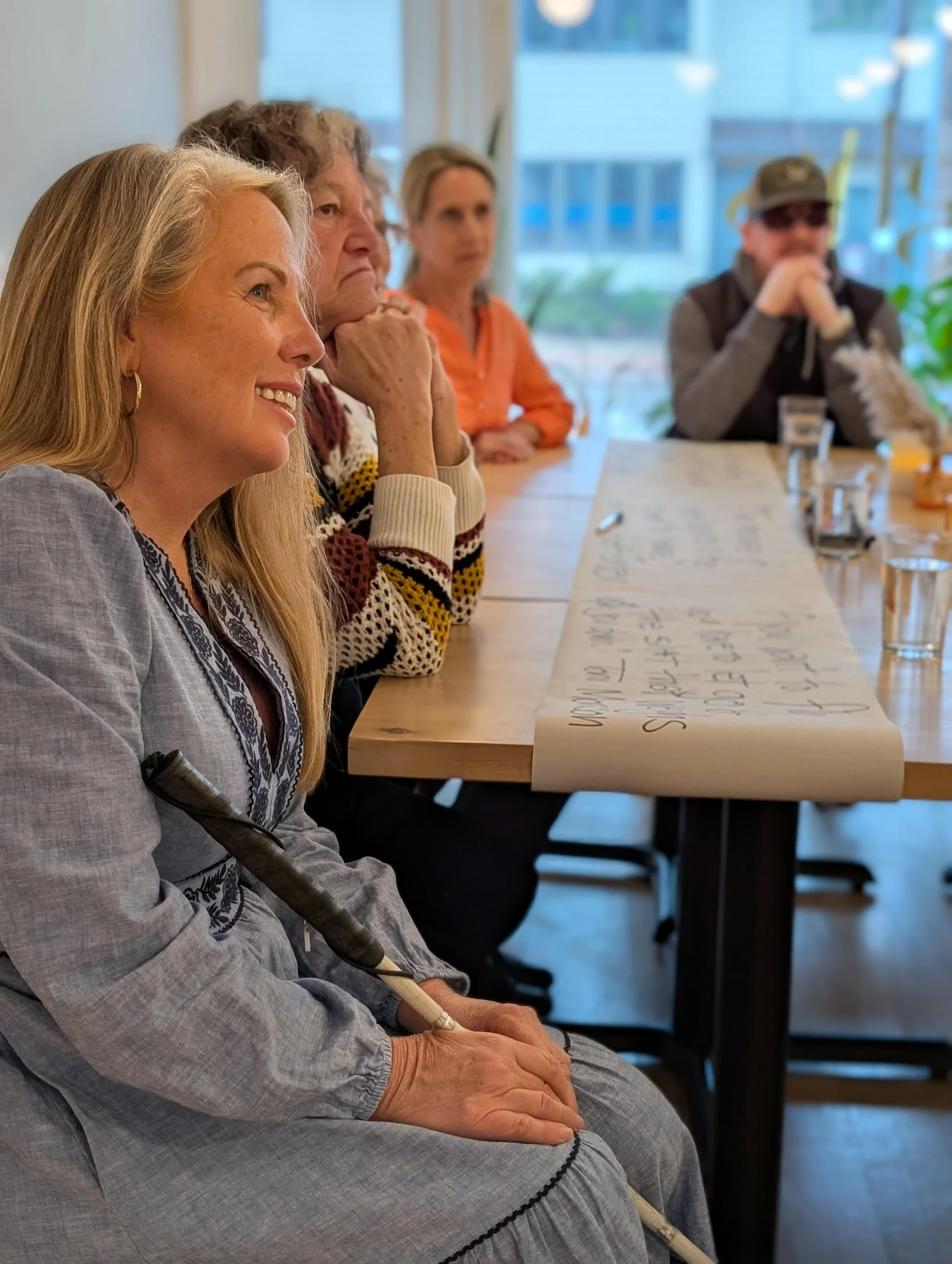 An indoor living room photograph showing Wendi Dwyer sitting in frame wearing a red cardigan and a bright turquoise ring. Her guide dog, Lily, is seen in the left hand corner of the image.